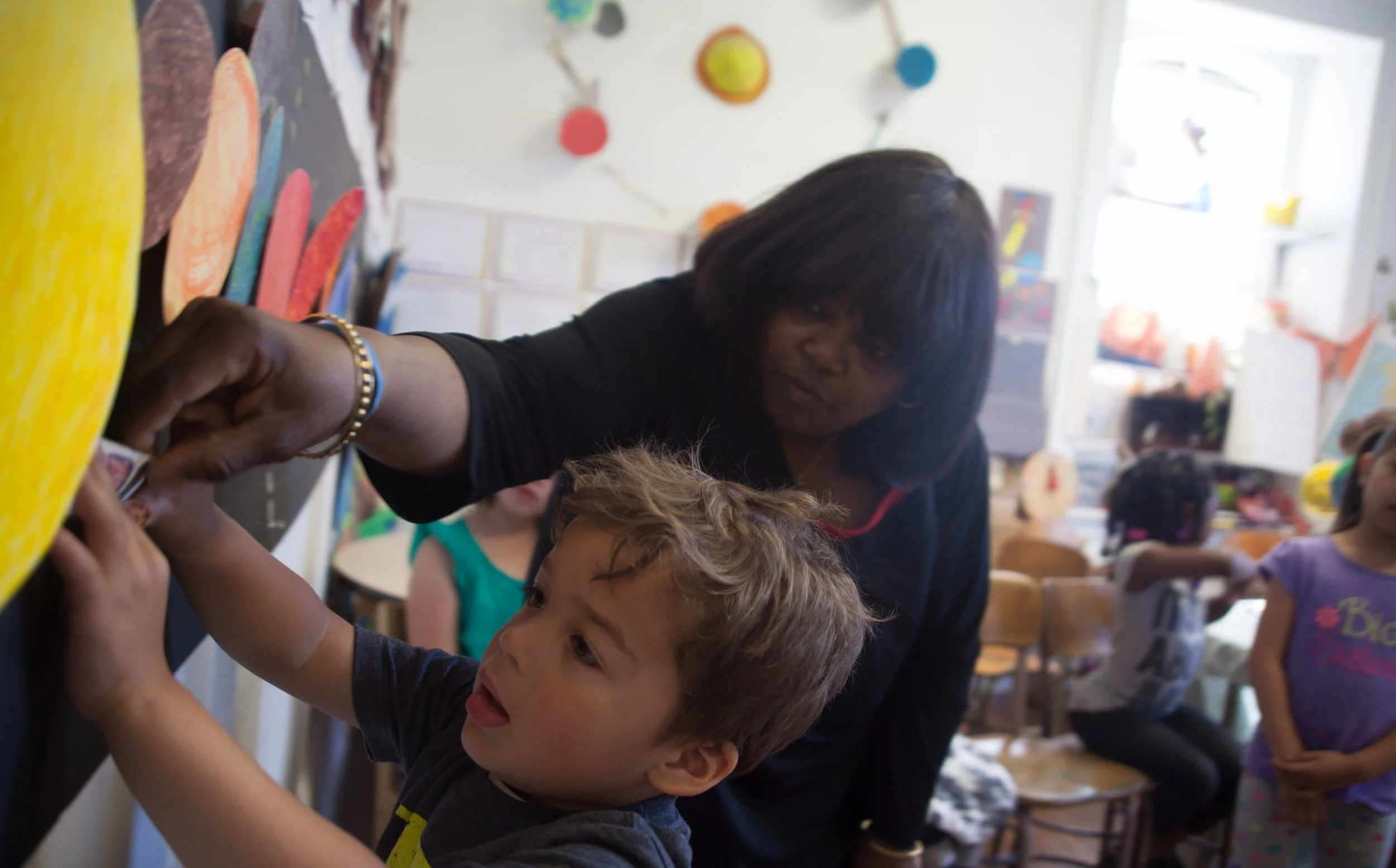 A teacher assisting a young boy with a classroom art project, with other children present in the background.
