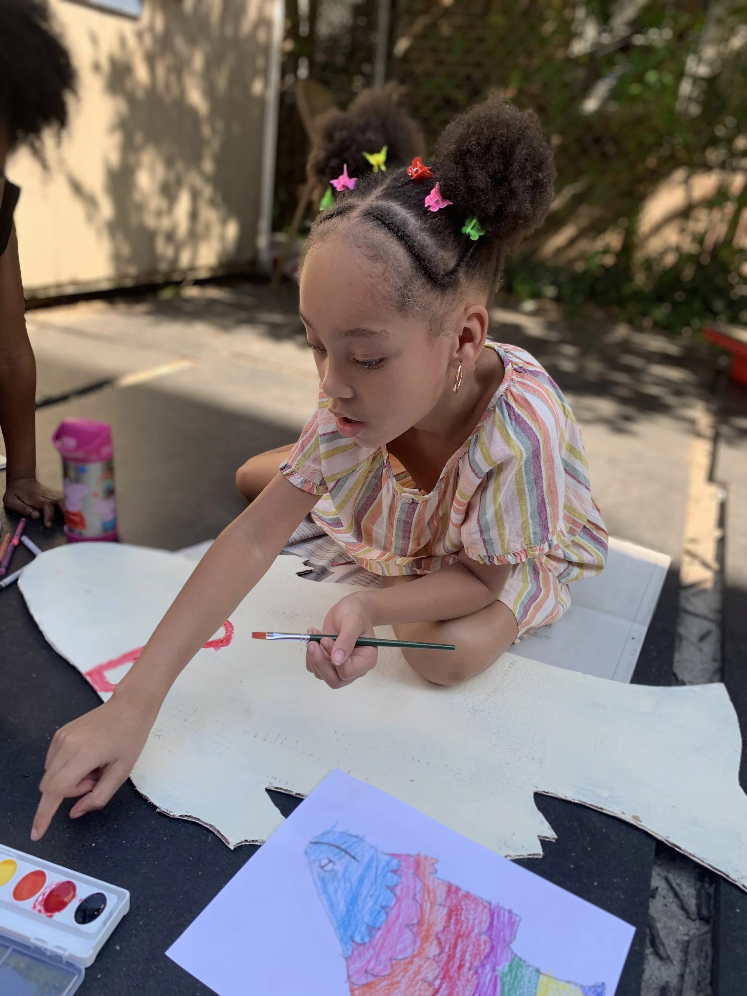 Young girl with braided hair decorated with colorful butterfly clips, sitting on a cardboard piece outdoors, painting on a piece of paper with watercolors and a paintbrush.