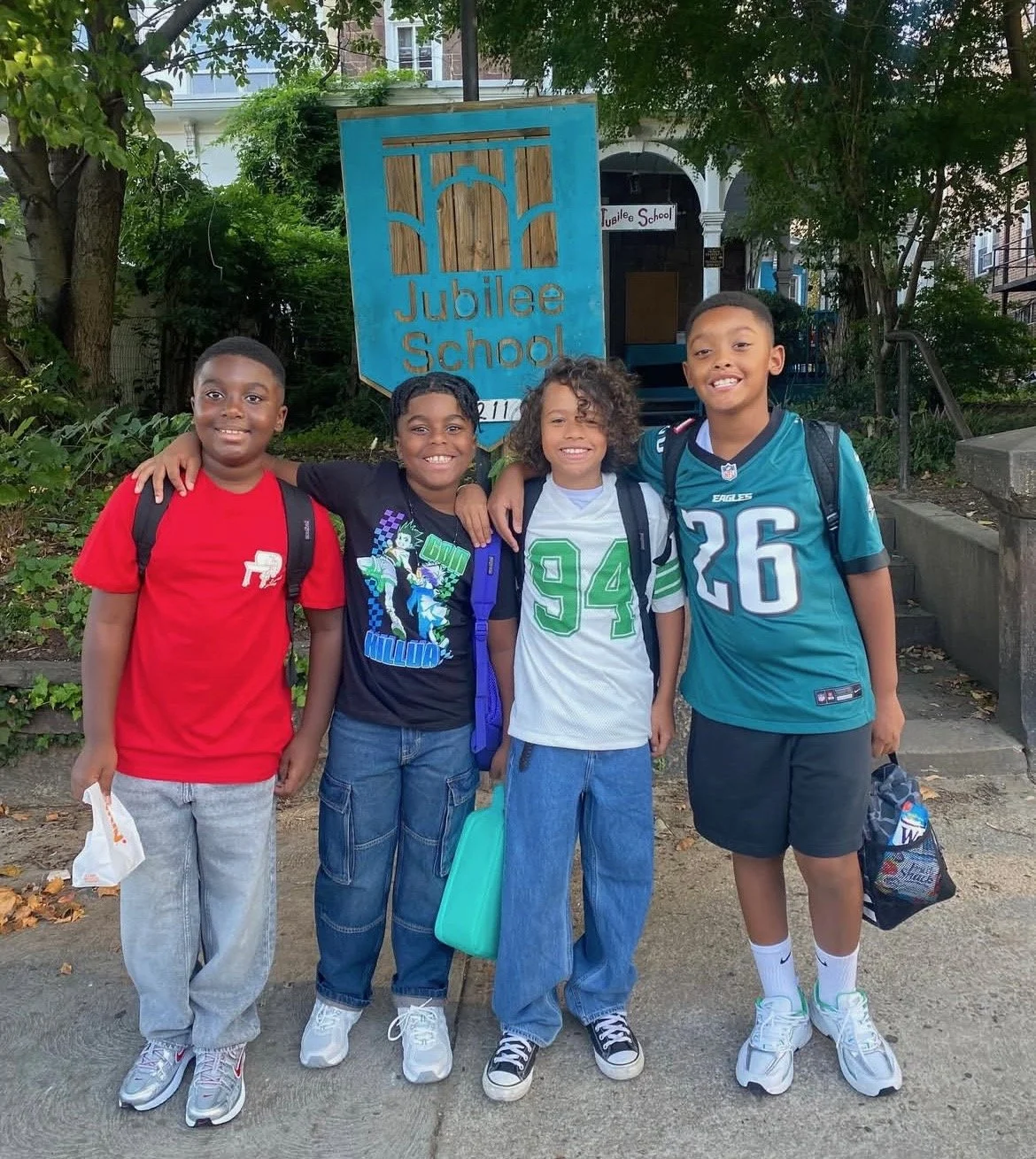 Four young boys standing in front of a blue sign that reads 'Jubilee School.' The boys are smiling, wearing backpacks, and standing on a sidewalk with trees and residential buildings in the background.