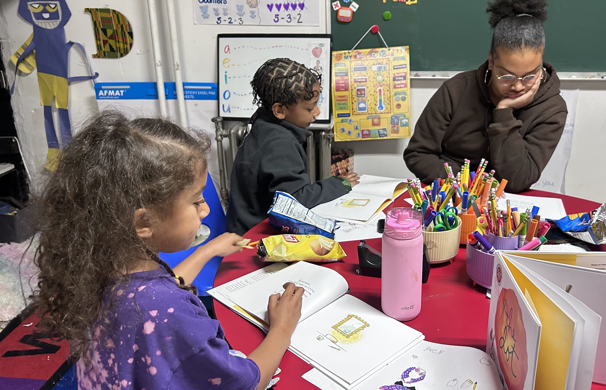 A classroom scene with three children sitting around a red table, engaged in reading and writing activities. The girl on the left has curly hair and is wearing a purple tie-dye shirt. The boy in the middle has braided hair and is focused on a book. The girl on the right wears glasses, has her hair in a bun, and is leaning on her hand. The table is filled with markers, pens, a pink water bottle, snacks, and open books. There are educational posters and a blackboard in the background.