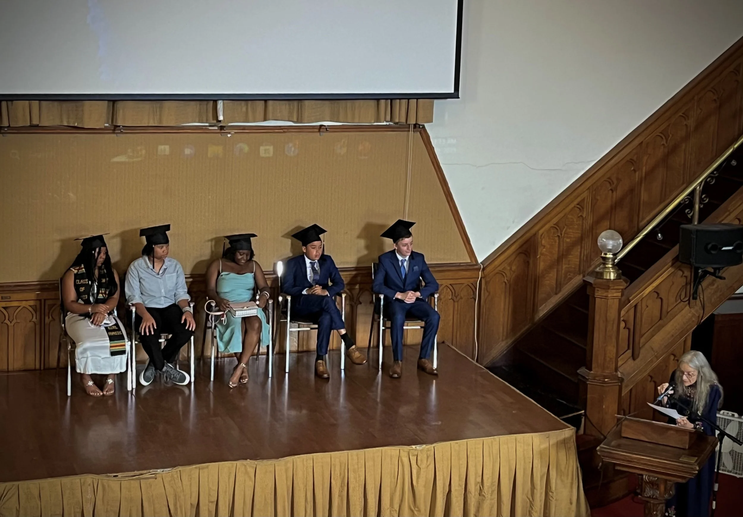 Six graduates wearing caps and gowns sitting on chairs on a stage, with a woman speaking at a podium to the right, and a large screen above them.