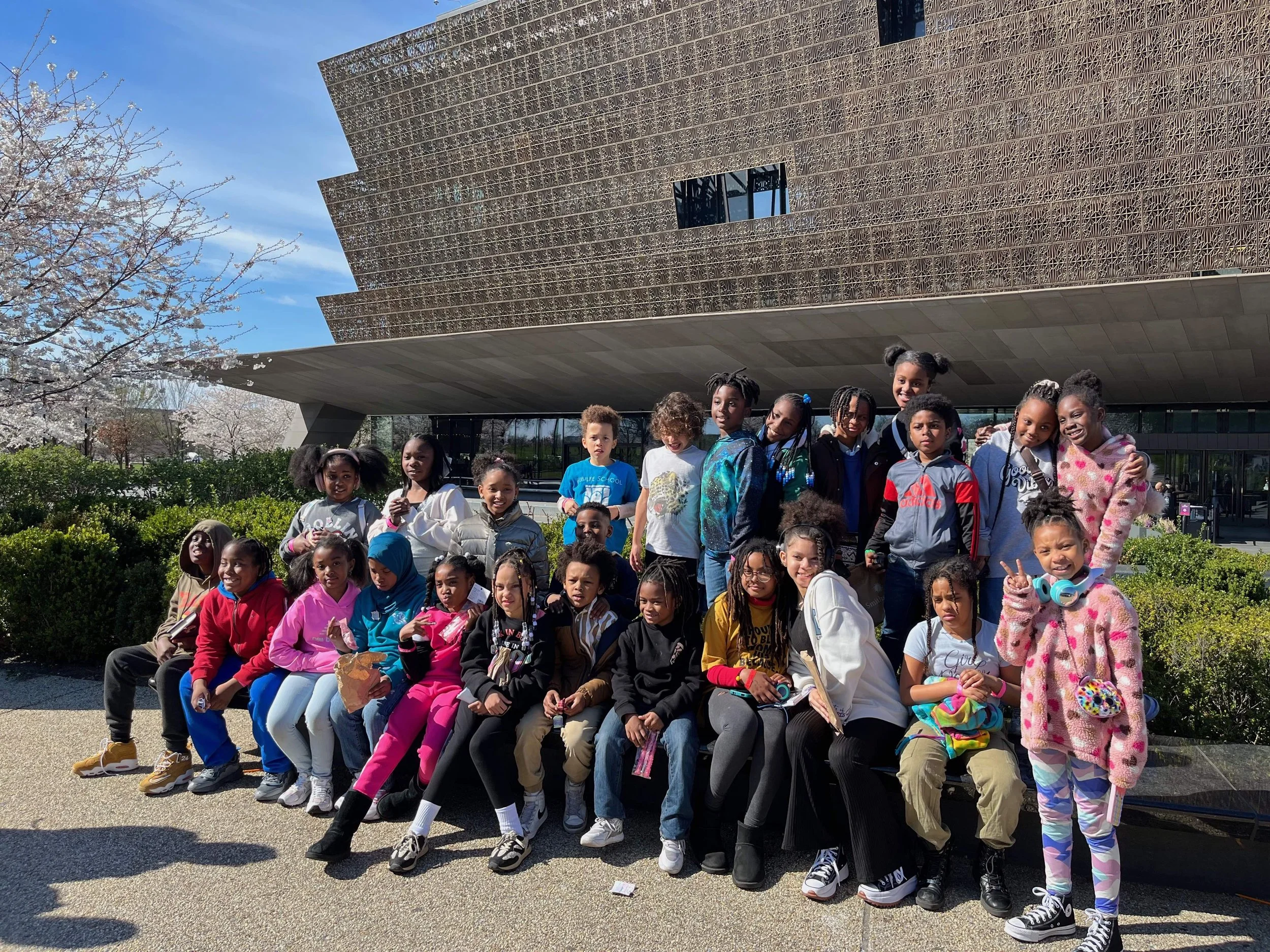 A group of children posing outdoors in front of a modern building with a brown, perforated facade. The children are sitting and standing on a park bench, some smiling and making peace signs. Spring blossoms are visible on trees in the background.