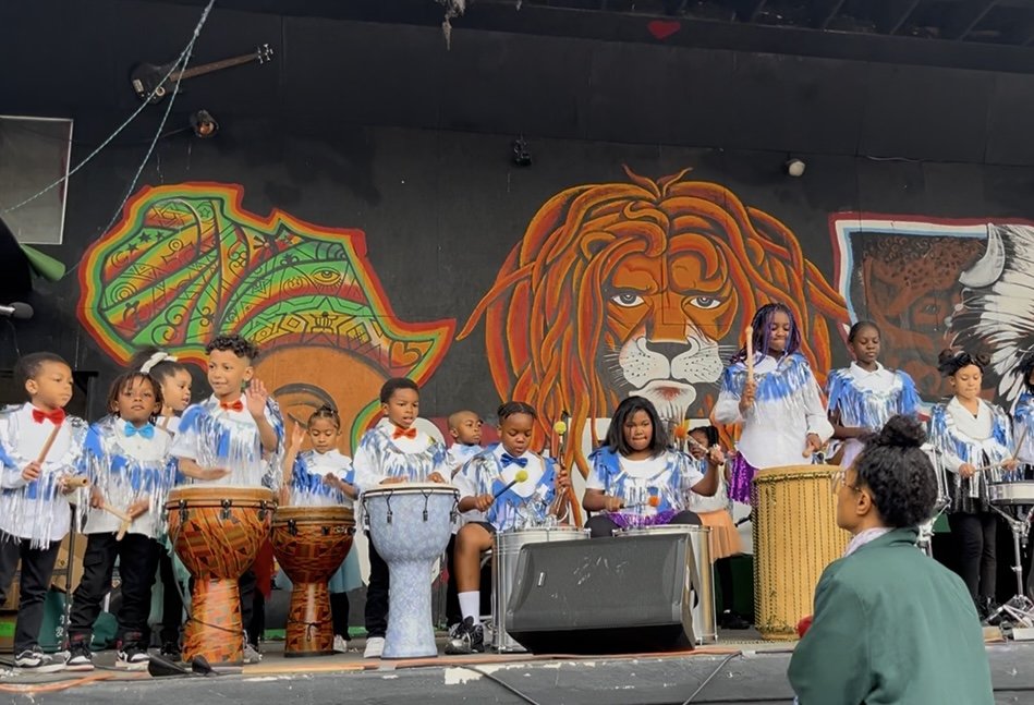 Group of children performing with drums and percussion instruments on stage, decorated with colorful African-themed murals, including a lion and an elephant.