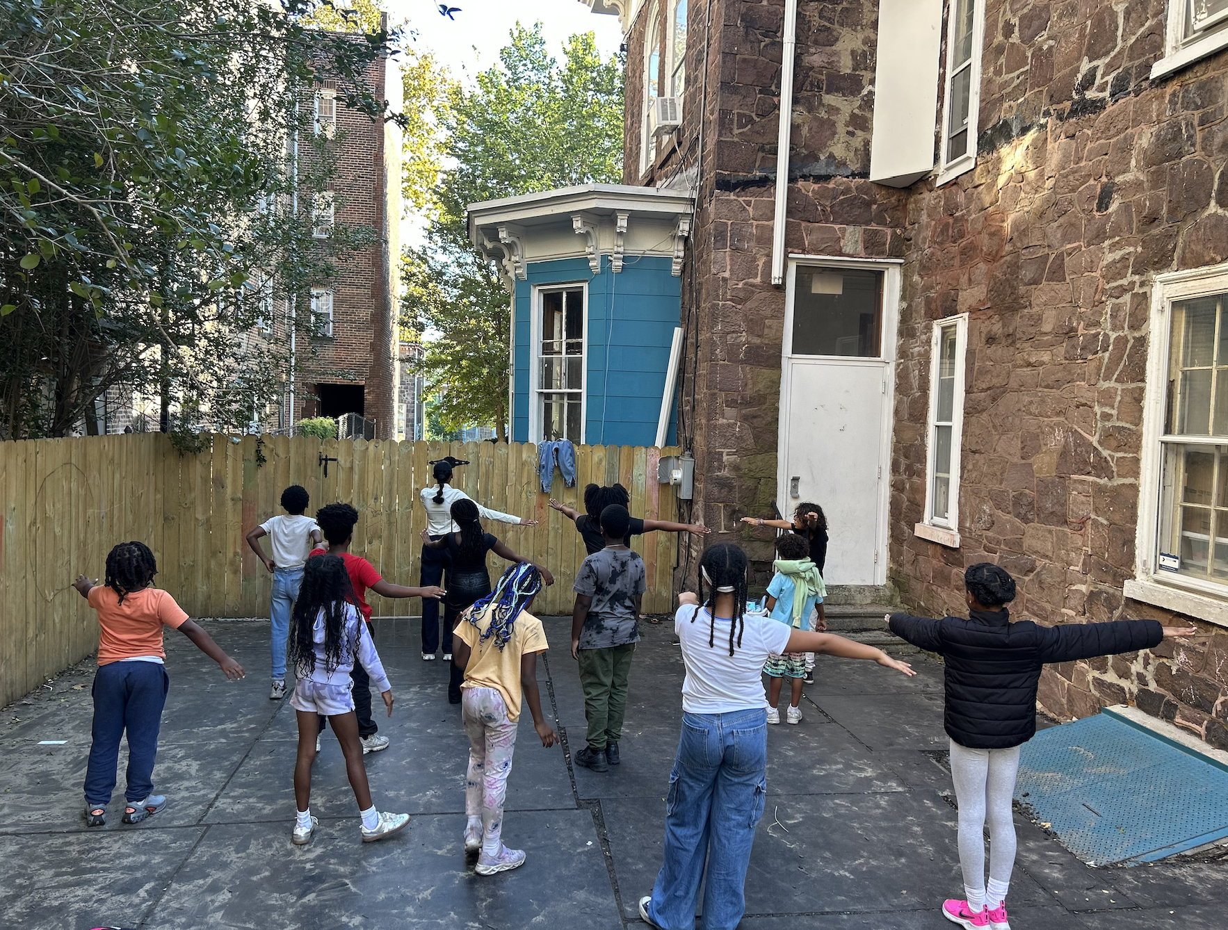 Children participating in an outdoor dance or exercise activity, extending arms outward, on a paved area next to a brick building with an adjacent blue house and a wooden fence.