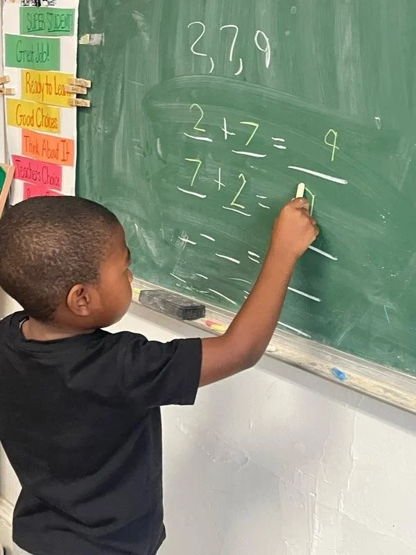 A young boy is solving math problems on a green chalkboard in a classroom. The problems include addition equations like 2 + 7 and 7 + 2. There are colorful signs with instructions or motivational messages on the wall to the left of the chalkboard.