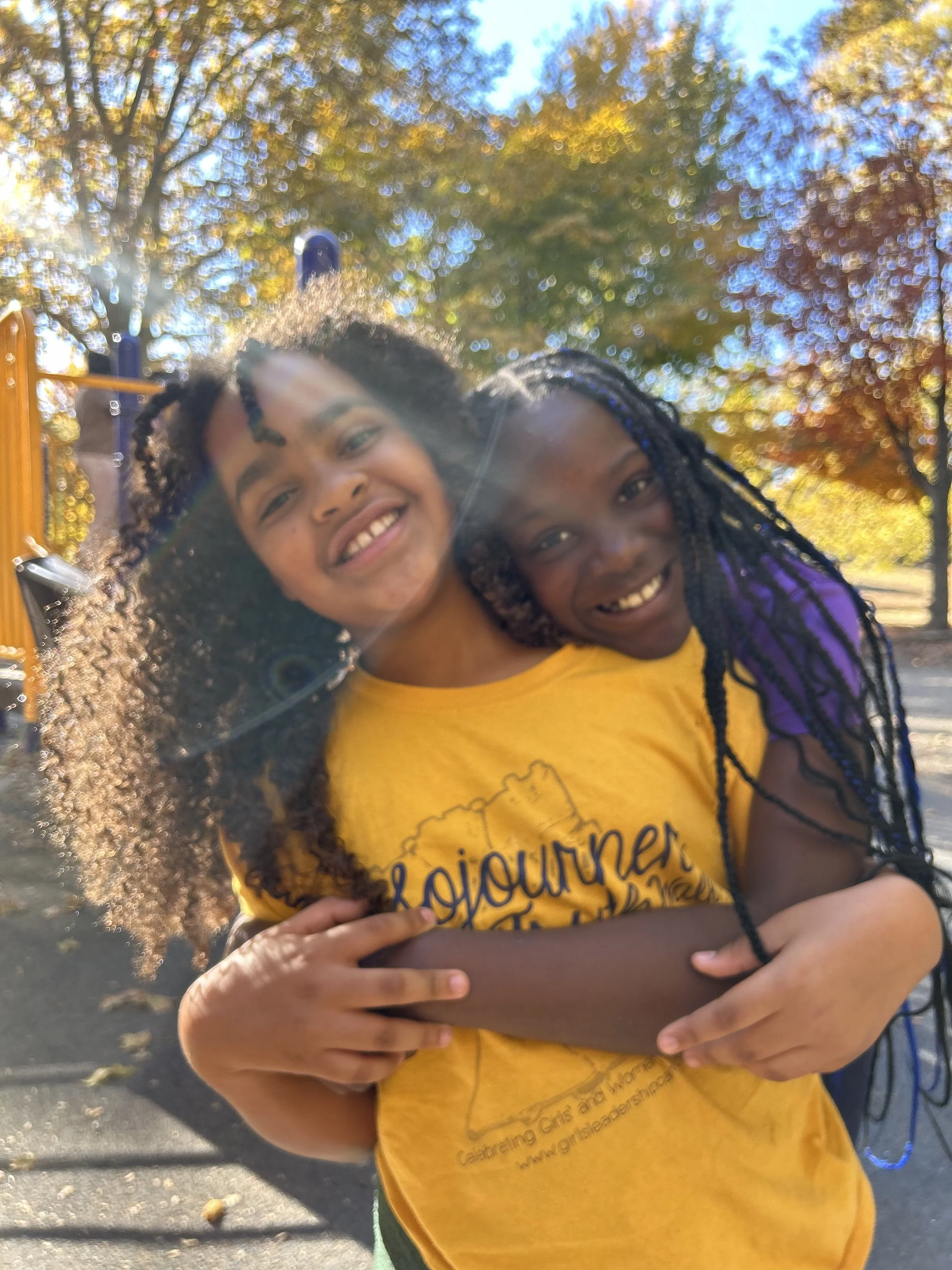 Two girls hugging and smiling outdoors on a sunny day, with autumn trees in the background.