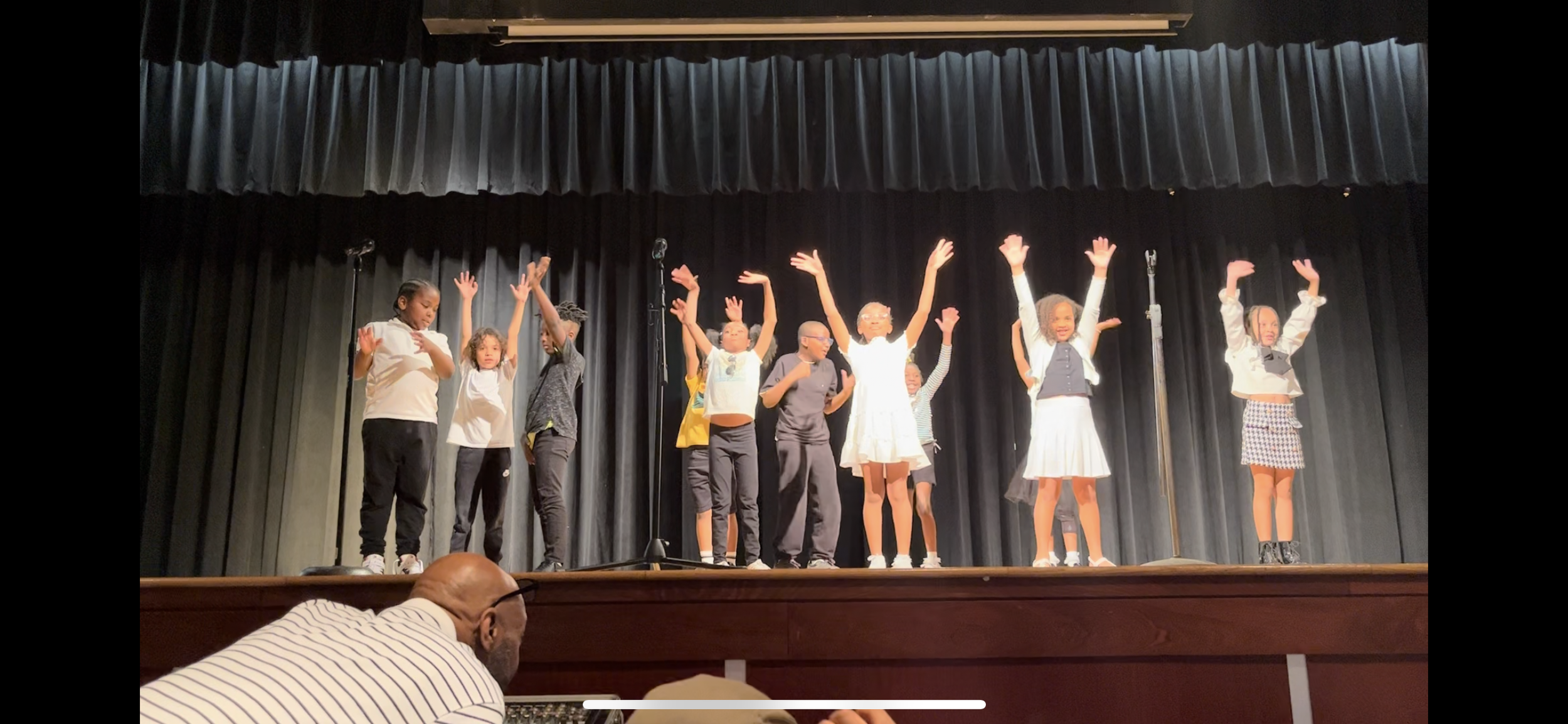 Children performing on stage with black curtains in the background, some with their hands raised, dressed in casual and school outfits.