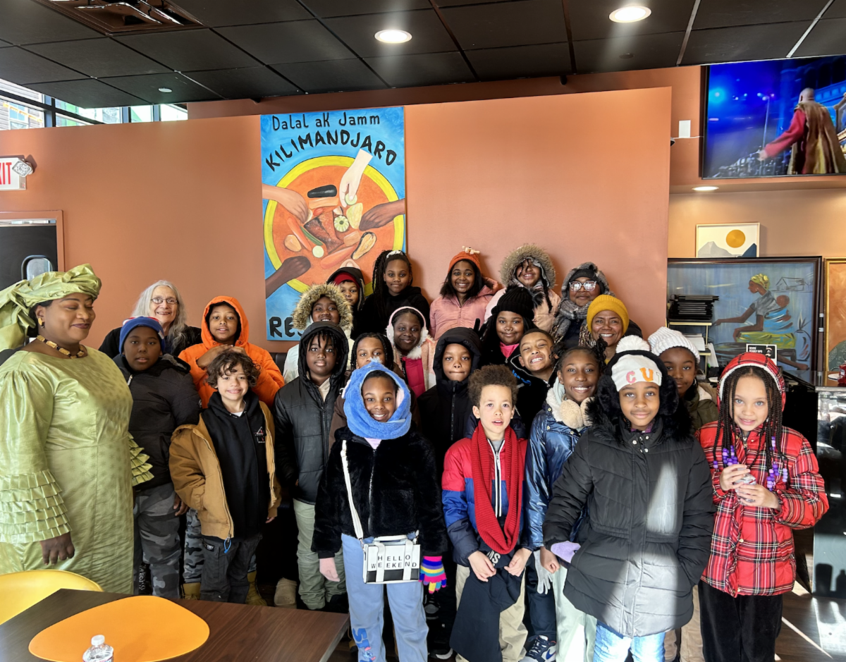 Group of children and adults posing for a photo indoors, some wearing jackets and winter clothing, with a woman in a green traditional outfit on the left, a colorful poster behind them, and television screens on the wall.