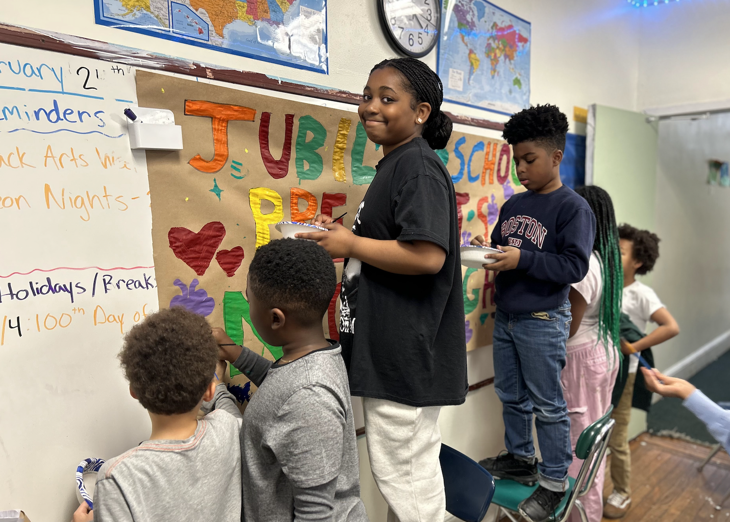 A group of children in a classroom participating in a celebration, with a colorful sign that says 'Jubilation &' and hearts on the wall.