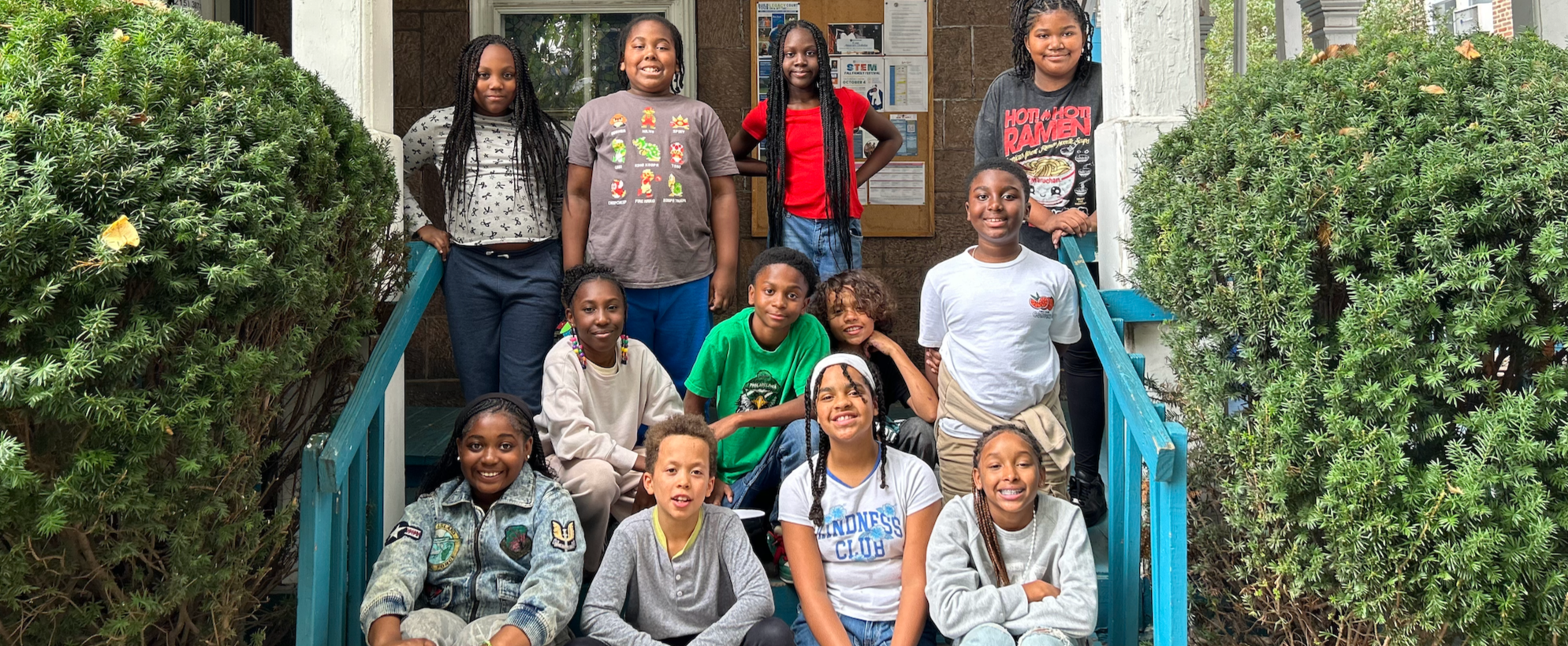 Group of 13 diverse children standing and sitting on the steps outside a building, smiling at the camera.