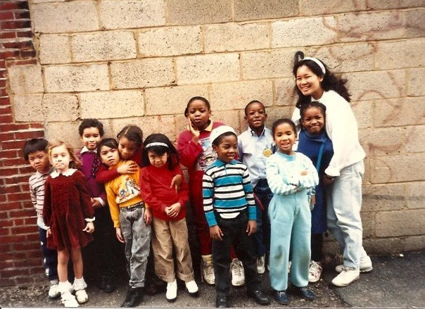 A group of young children standing next to a smiling woman in front of a brick wall.