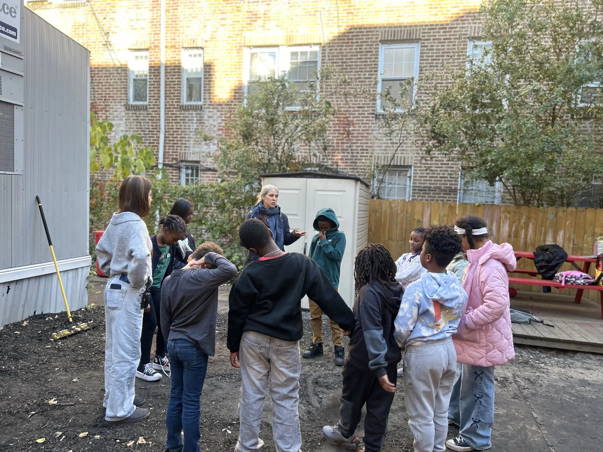 A group of children and a woman are standing in a circle outside in a yard, engaging in a discussion or activity. The yard has a brick building in the background, some trees, and a small shed. Some children are wearing hoodies and sweatpants, and one child is wearing a pink coat. There are gardening tools like a rake leaning against a wall.