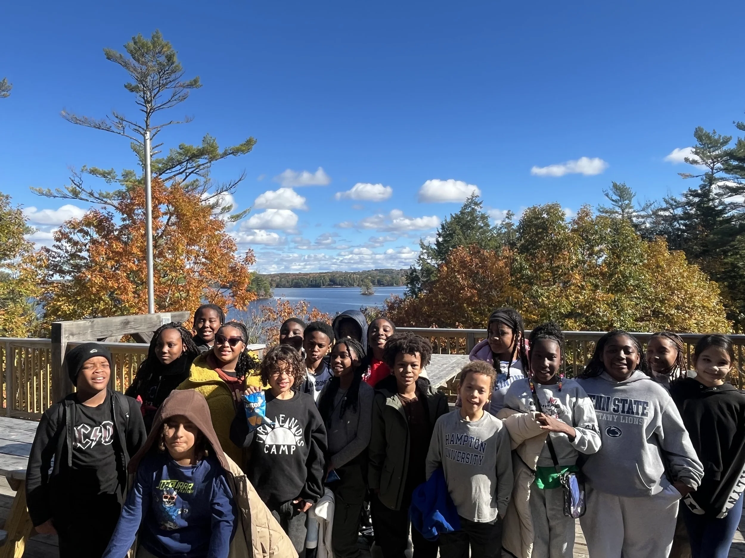 Group of children and teenagers outdoors on a wooden deck with autumn trees and a lake in the background under a blue sky with scattered clouds.