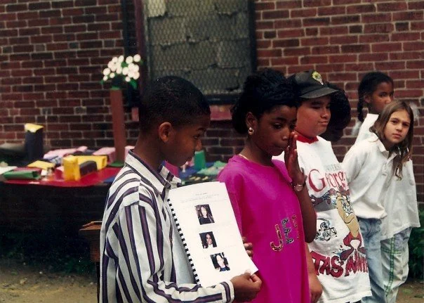 Five children standing in a row outdoors, with a brick wall and a table with colorful items and flowers in the background.