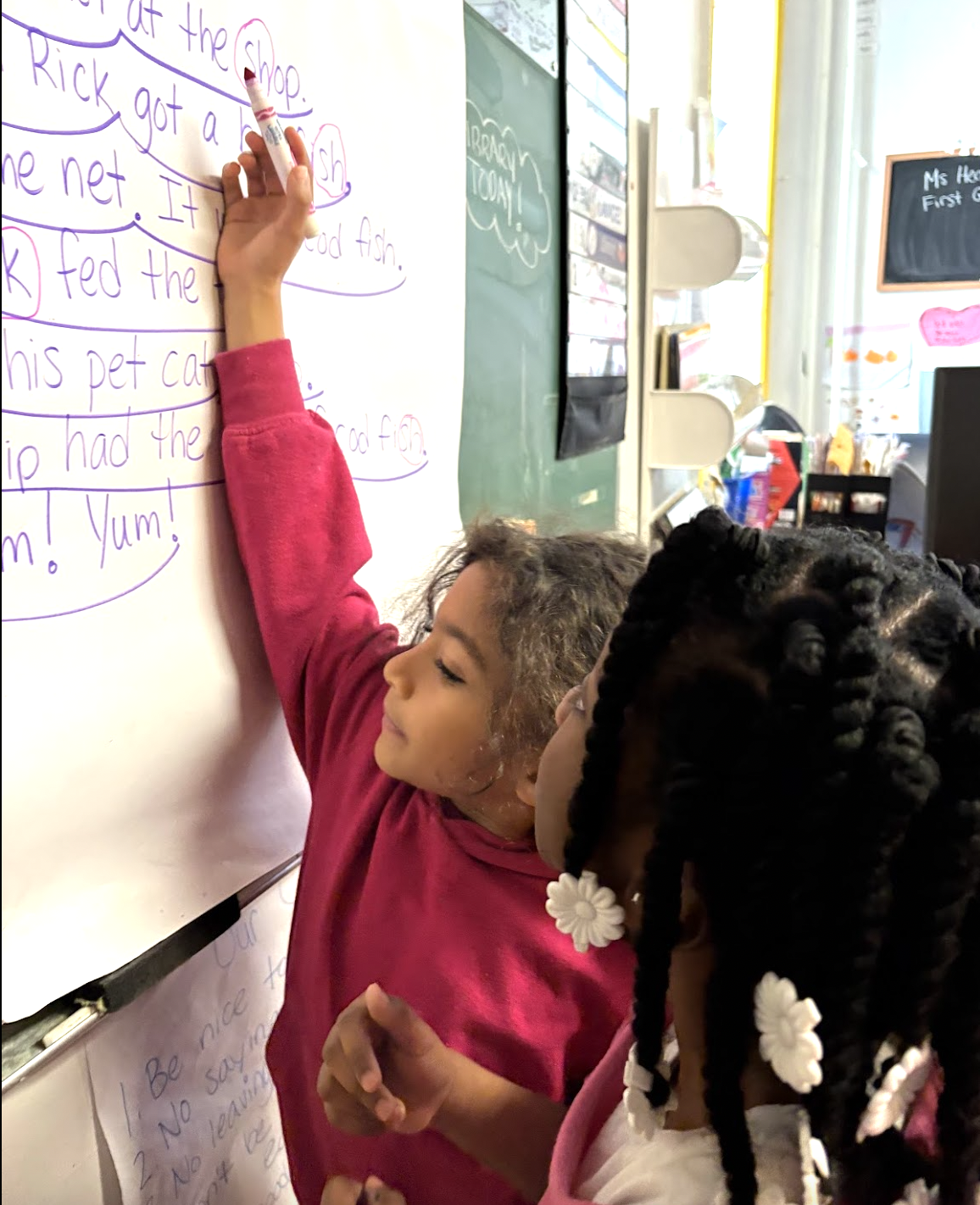 Two young girls are writing on a whiteboard in a classroom. One girl is wearing a pink long-sleeve shirt and is using a marker to write, while the other girl with braids decorated with white flowers is observing.