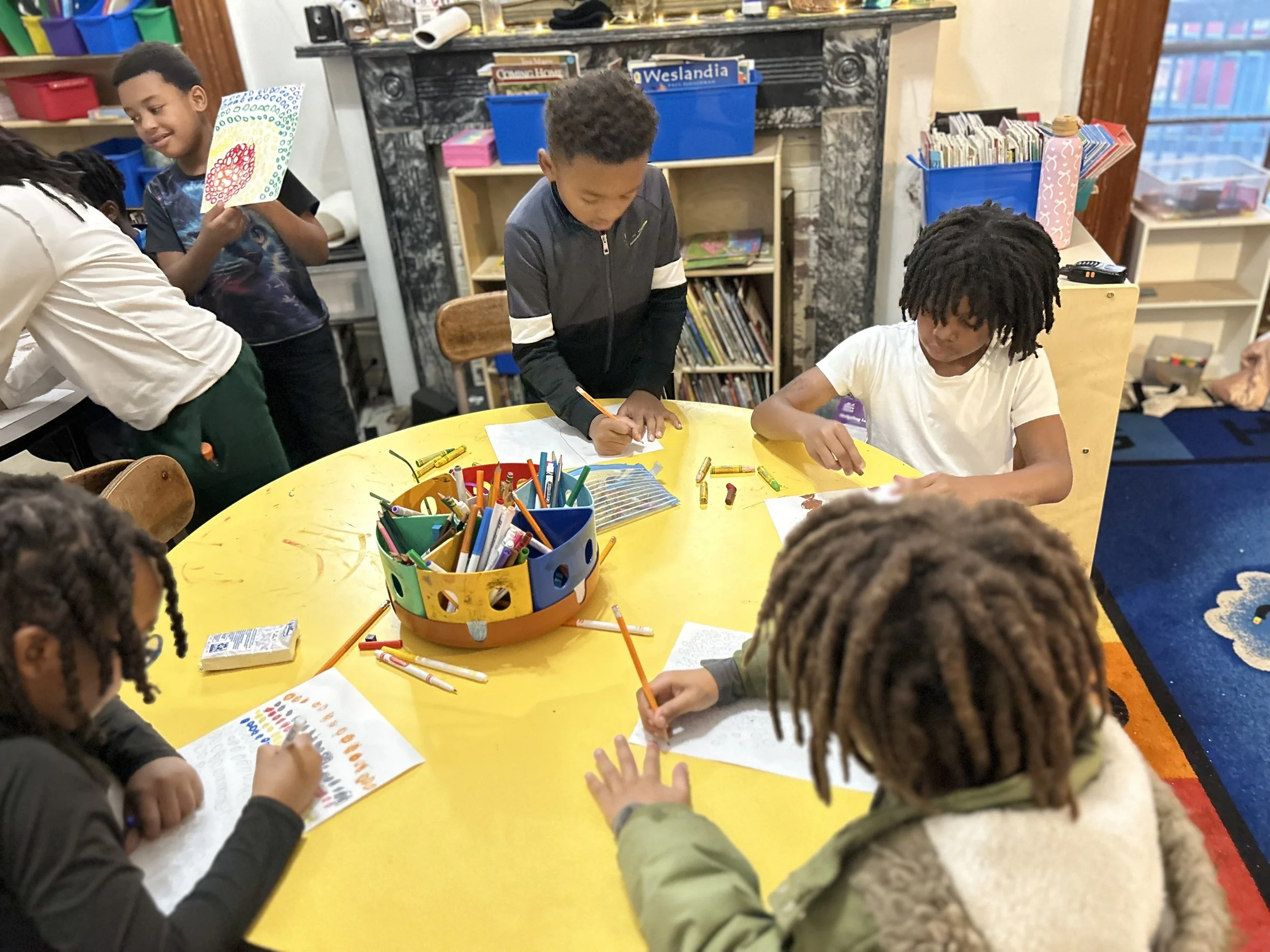 Children drawing and coloring at a yellow table in a classroom, with art supplies in a colorful container in the center.