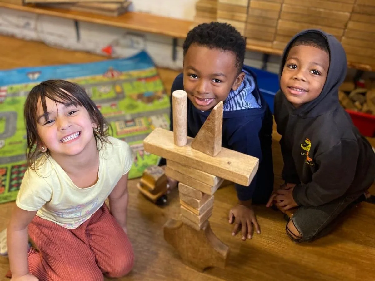 Three children smiling and playing with a tower of wooden blocks indoors.