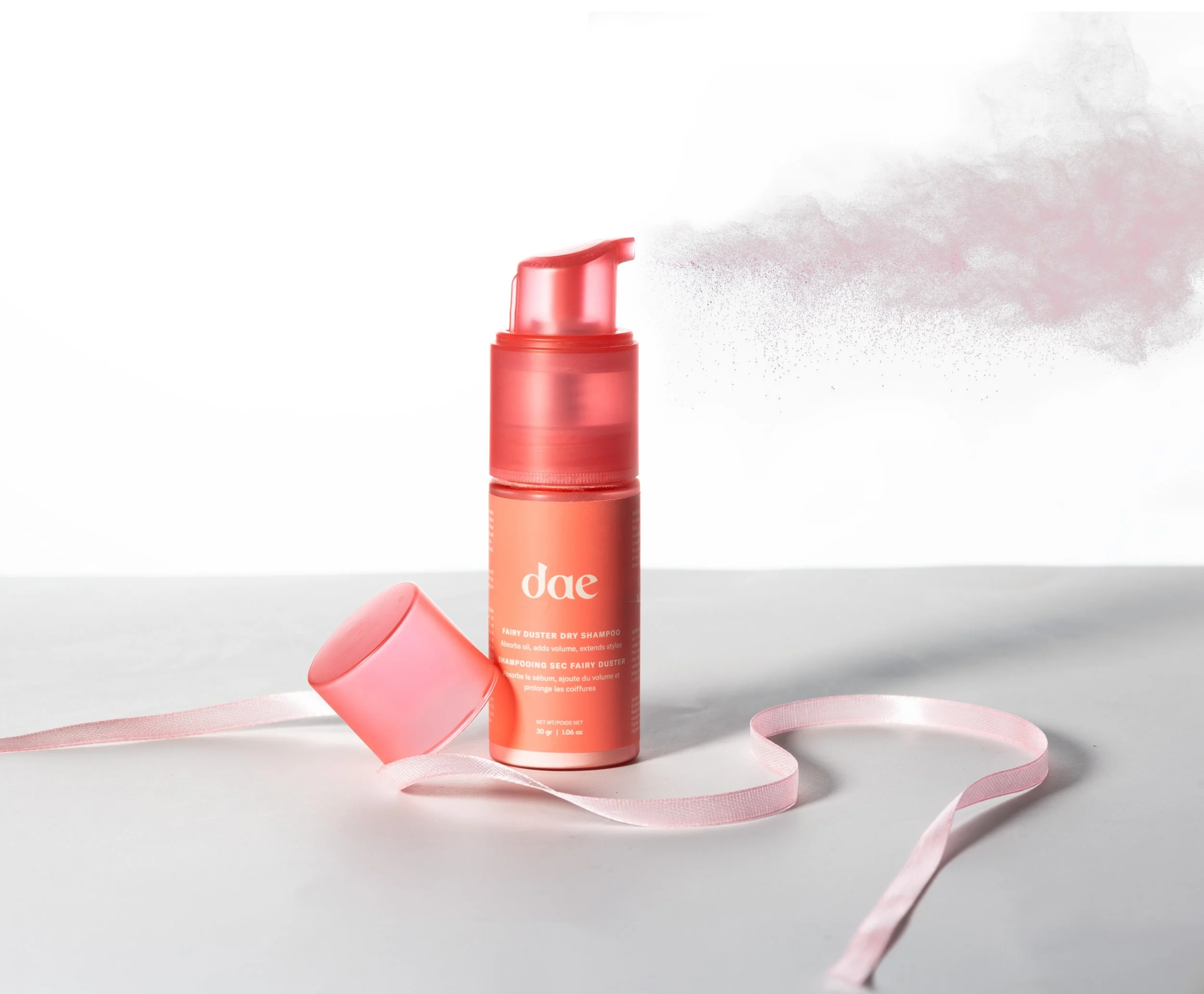 A coral-colored bottle of dry shampoo with a pink ribbon and spray of powder coming out of the nozzle.