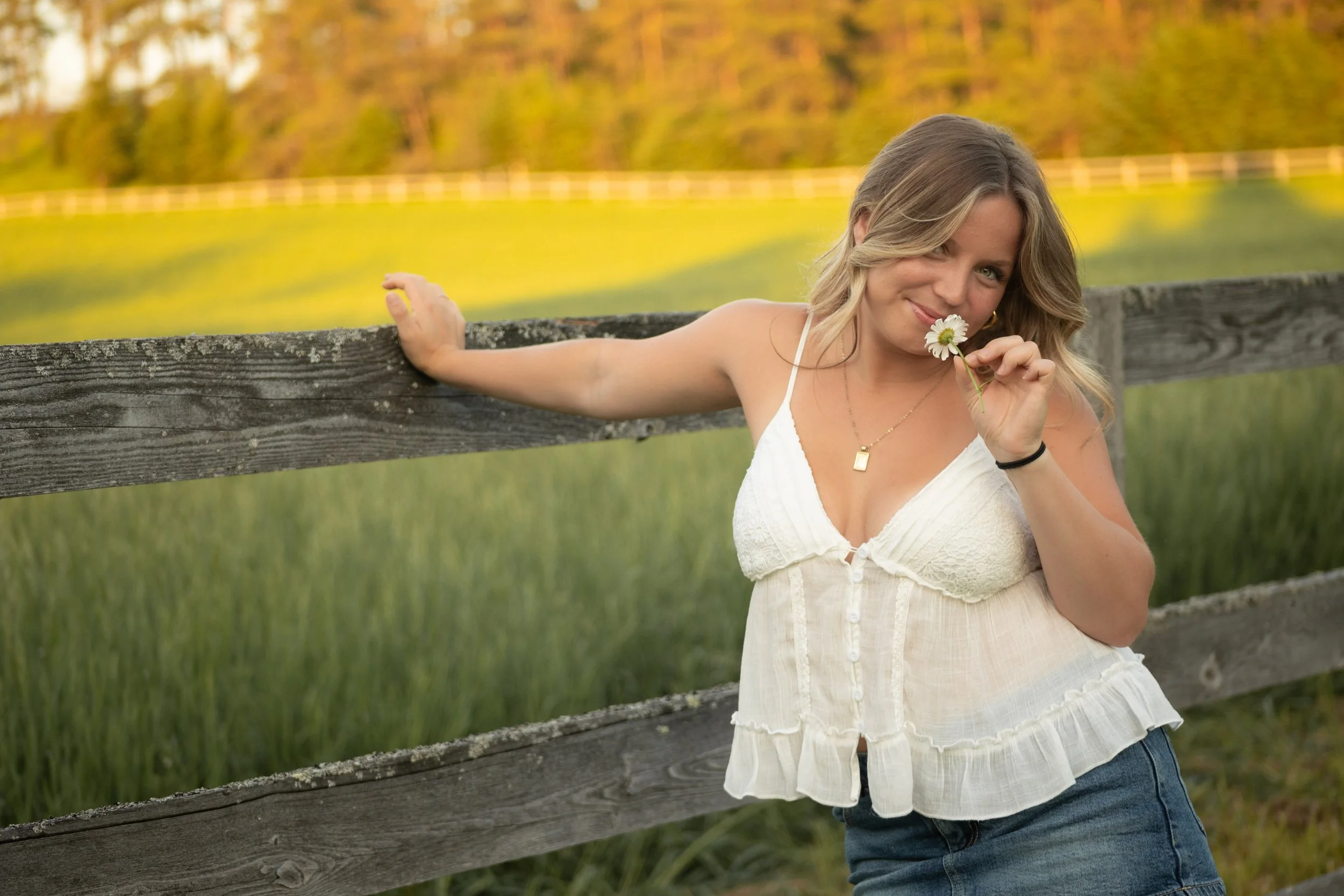 A woman with long, wavy blonde hair, wearing a white sleeveless top and blue jeans, smiling and holding a daisy flower near her face while leaning against a wooden fence in a grassy field with trees in the background during sunset.