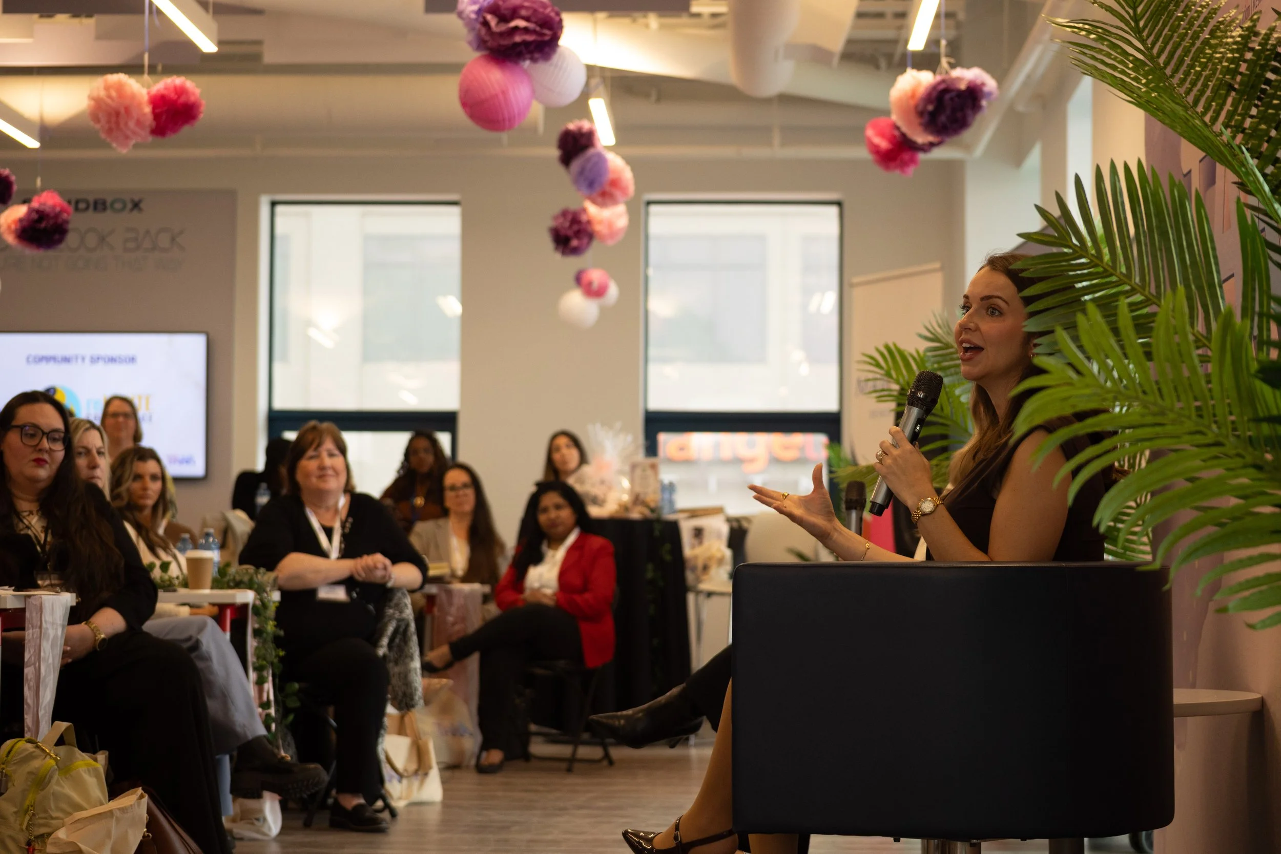 A woman holding a microphone speaking to an audience at an indoor event decorated with pink, purple, and white hanging paper lanterns and flowers. Several women are seated listening attentively.