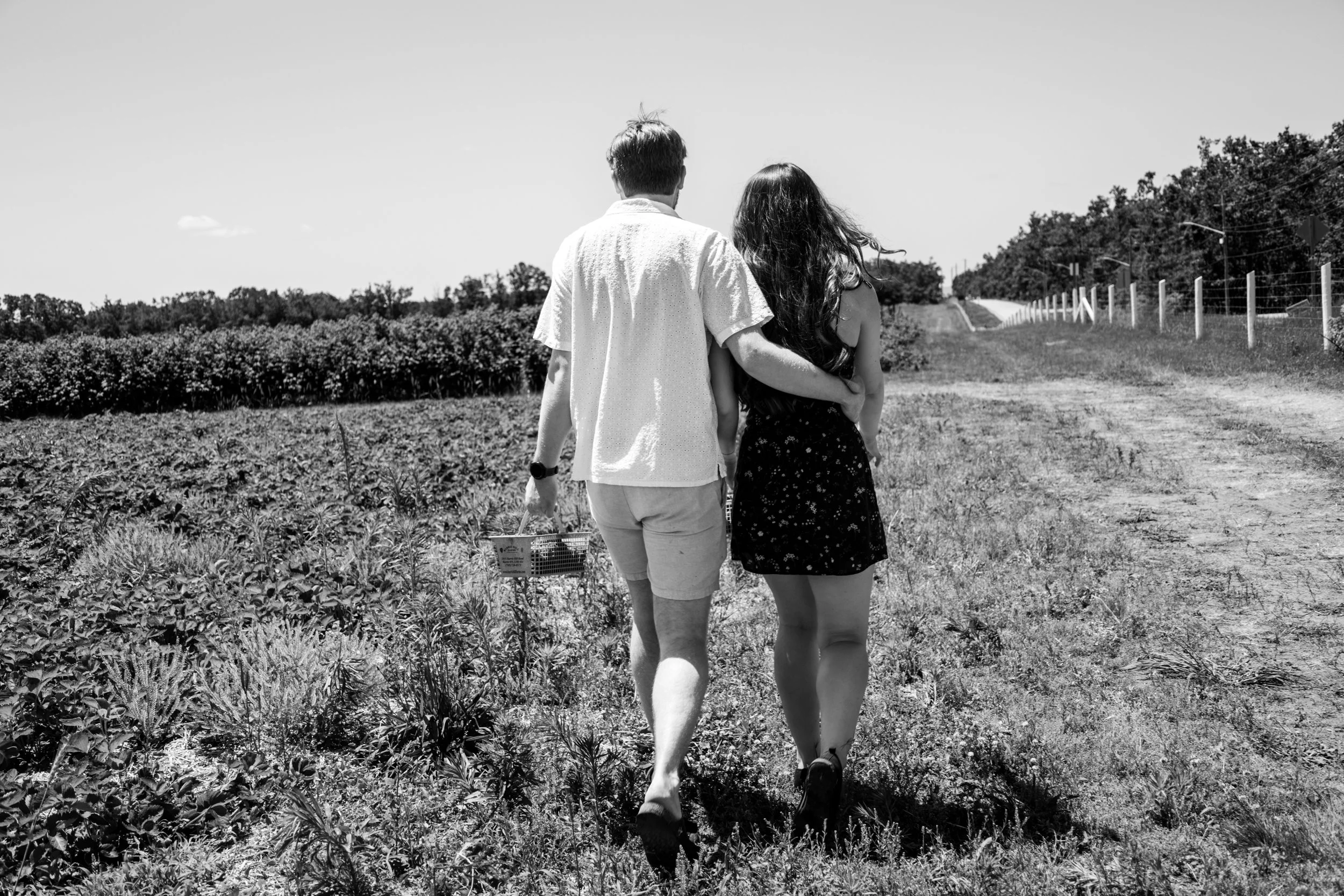 A black and white photo of a young man and woman walking through a farm or garden, with the man holding a basket and they are seen from behind.