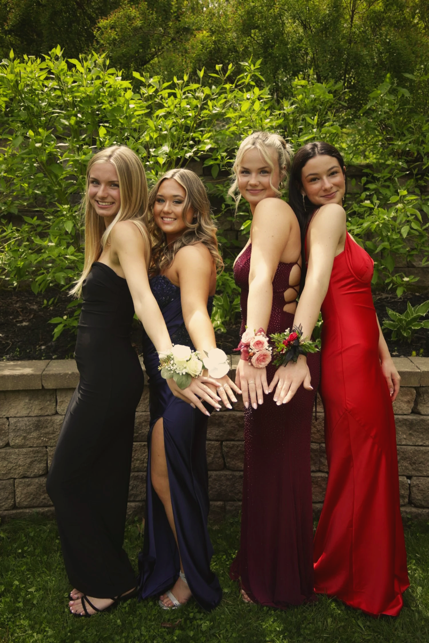Four young women in formal dresses pose outdoors in front of a green leafy hedge, smiling and holding flowers, with one wearing a black dress, one in a dark blue dress, one in a burgundy dress, and one in a red dress.