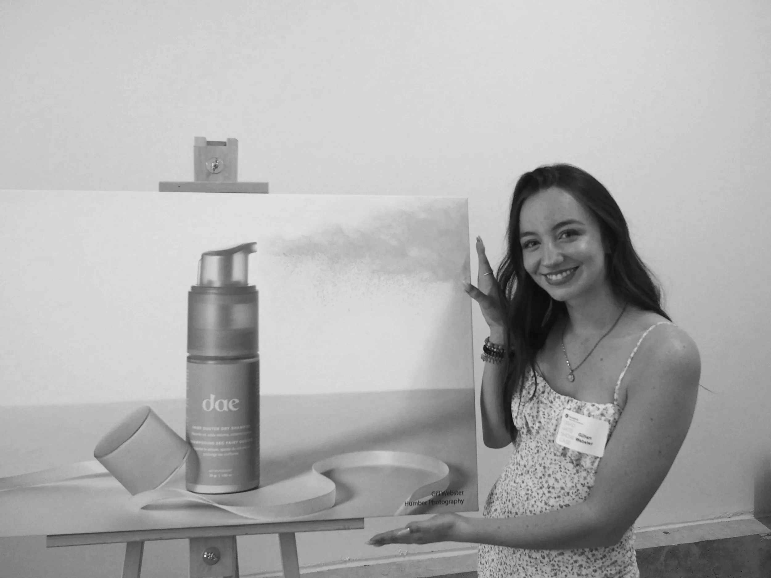 A woman smiling and pointing at a poster display with a picture of a spray bottle and text that reads 'dae' and 'THE BETTER SPRAY SPLASH.' She is wearing a patterned dress and has a name tag on her chest.
