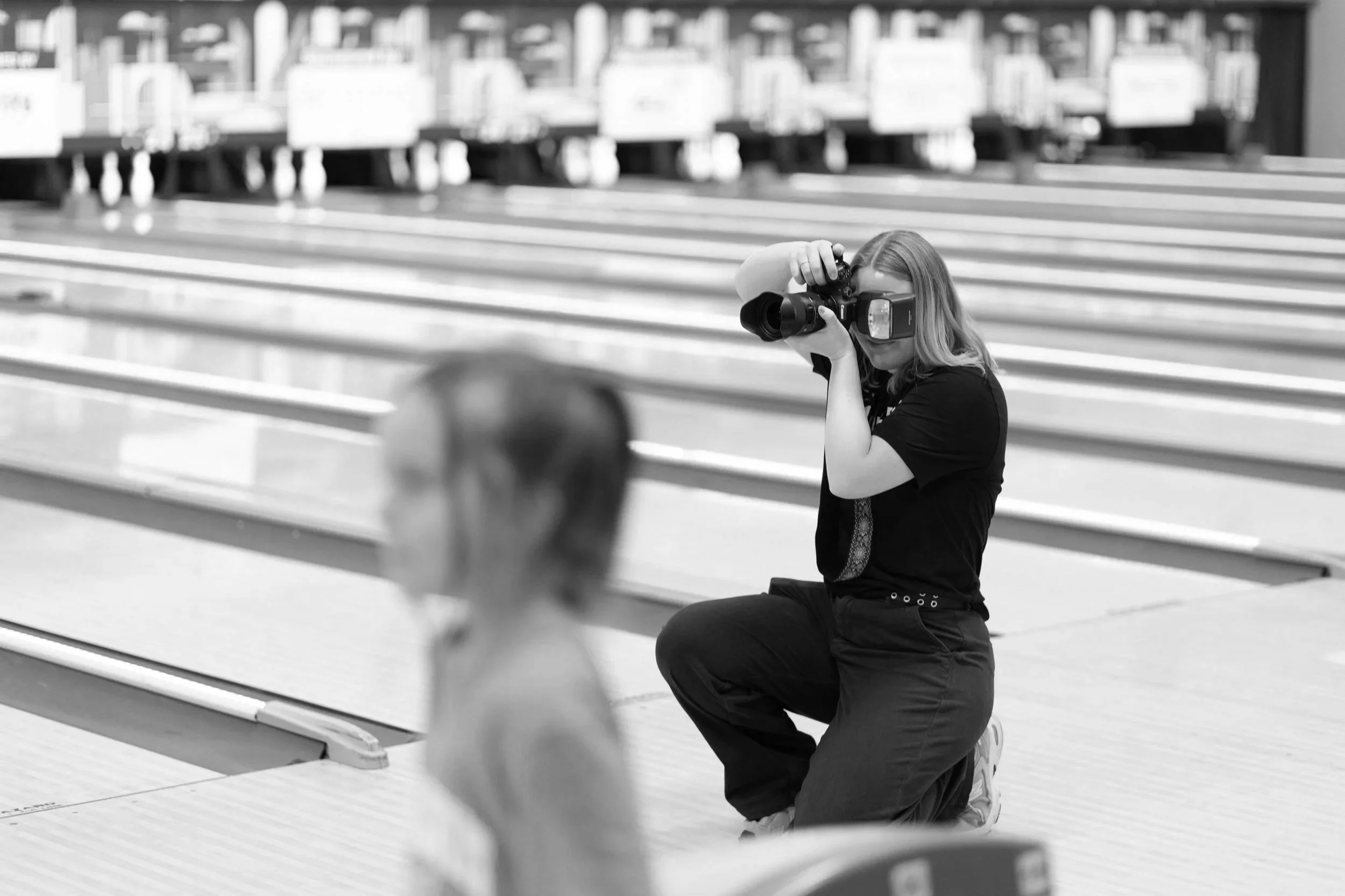 A woman with long hair kneeling on one knee at a bowling alley, aiming a camera towards the bowling pins and lanes in the background.
