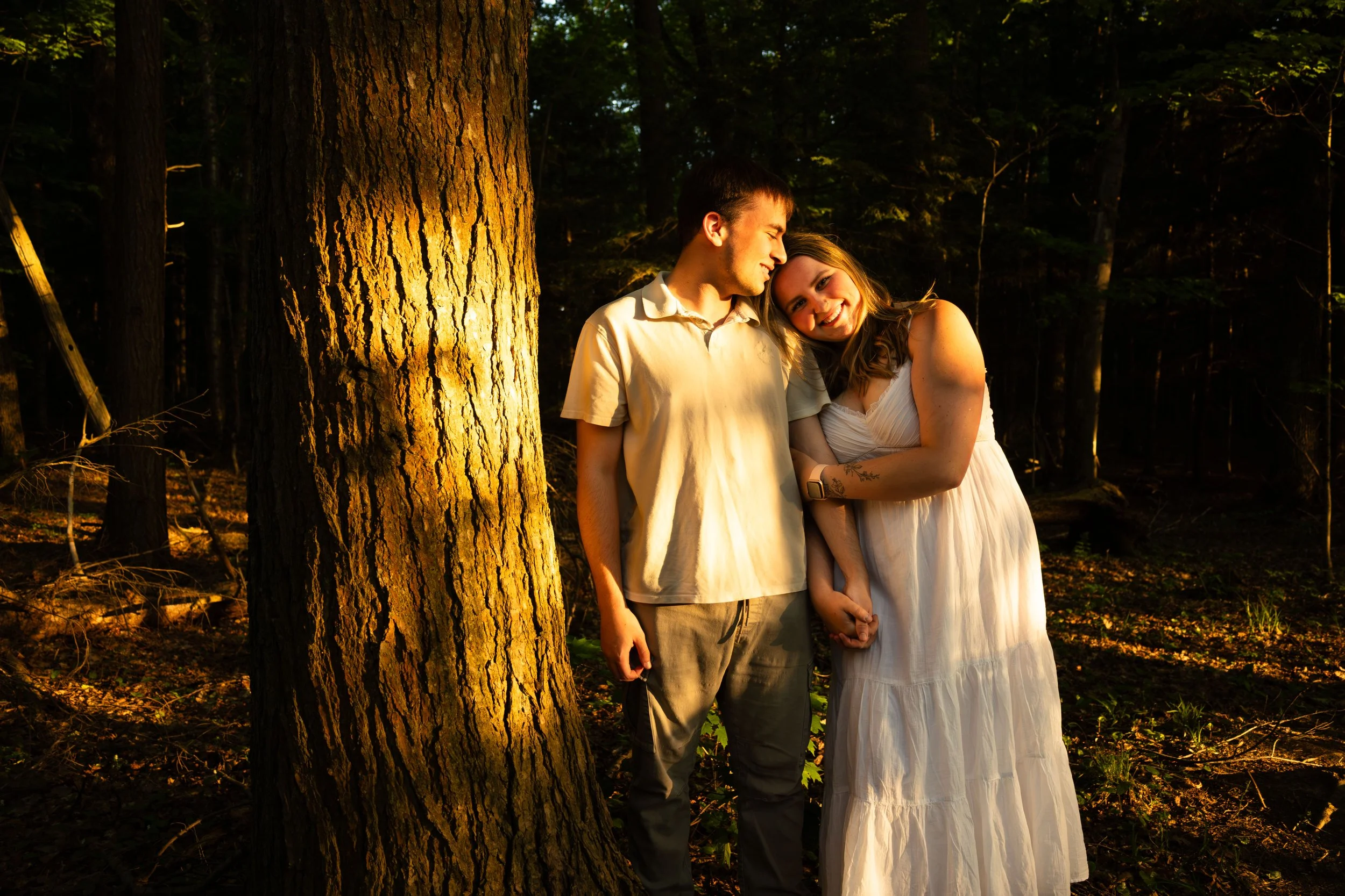 A young couple standing next to a large tree in a forest at sunset, smiling and embracing.