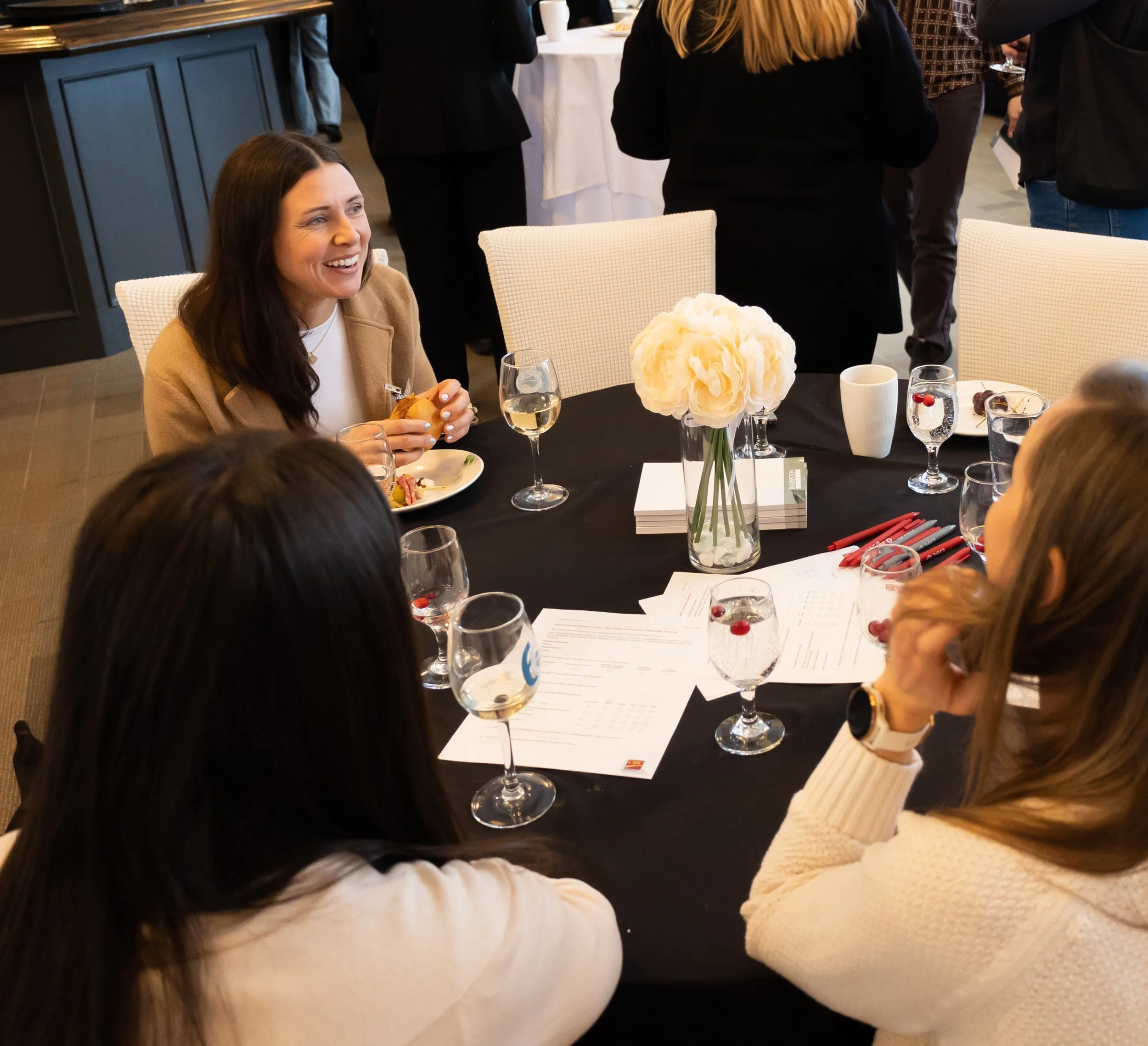 A woman smiling, sitting at a table with food and drinks, at a social gathering or celebration, with other people standing and talking in the background.