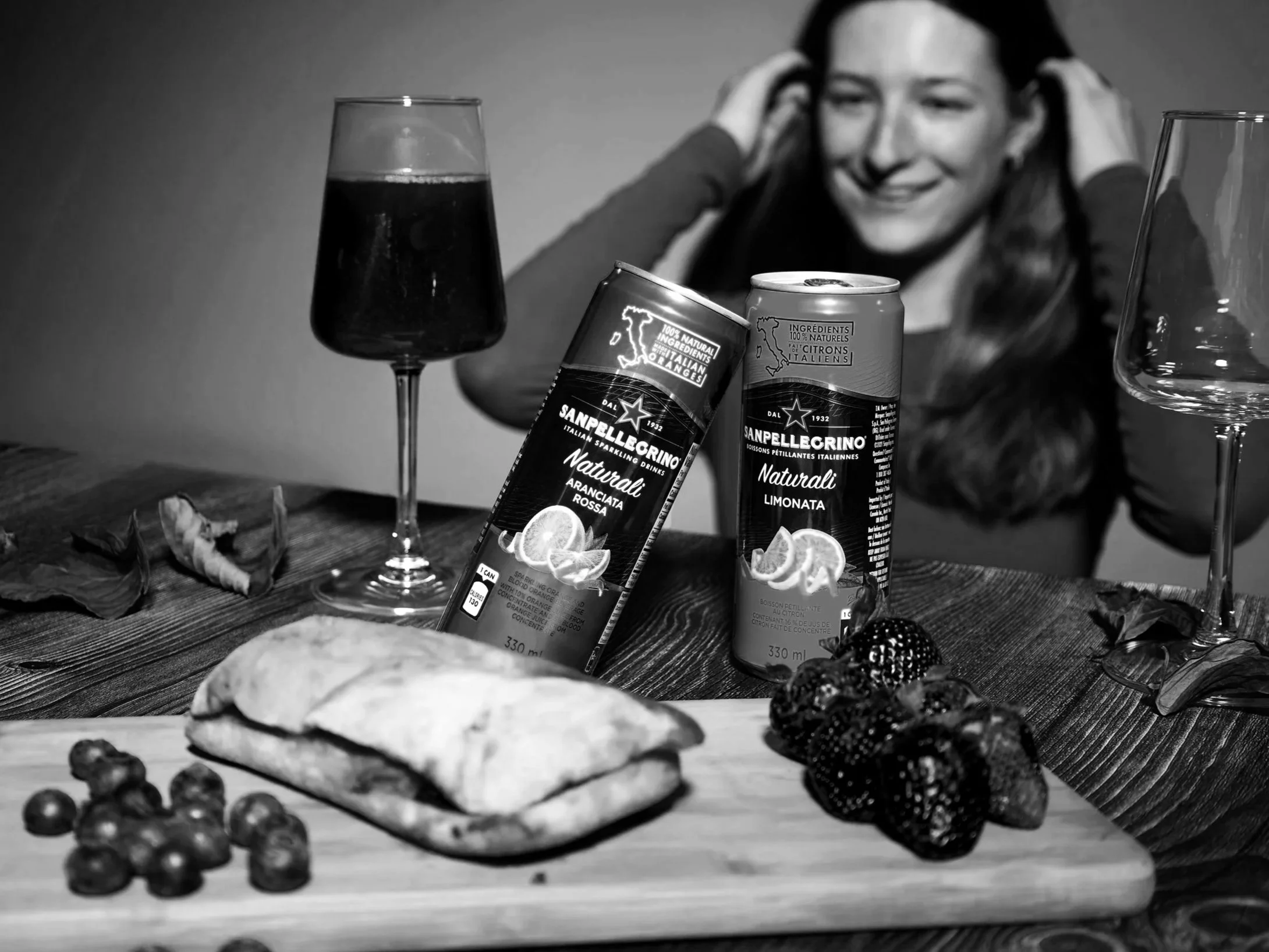 A woman smiling with her hands on her head sitting at a table with two cans of Sanpellegrino soda, a glass of dark beverage, a pizza, and various berries and blackberries.