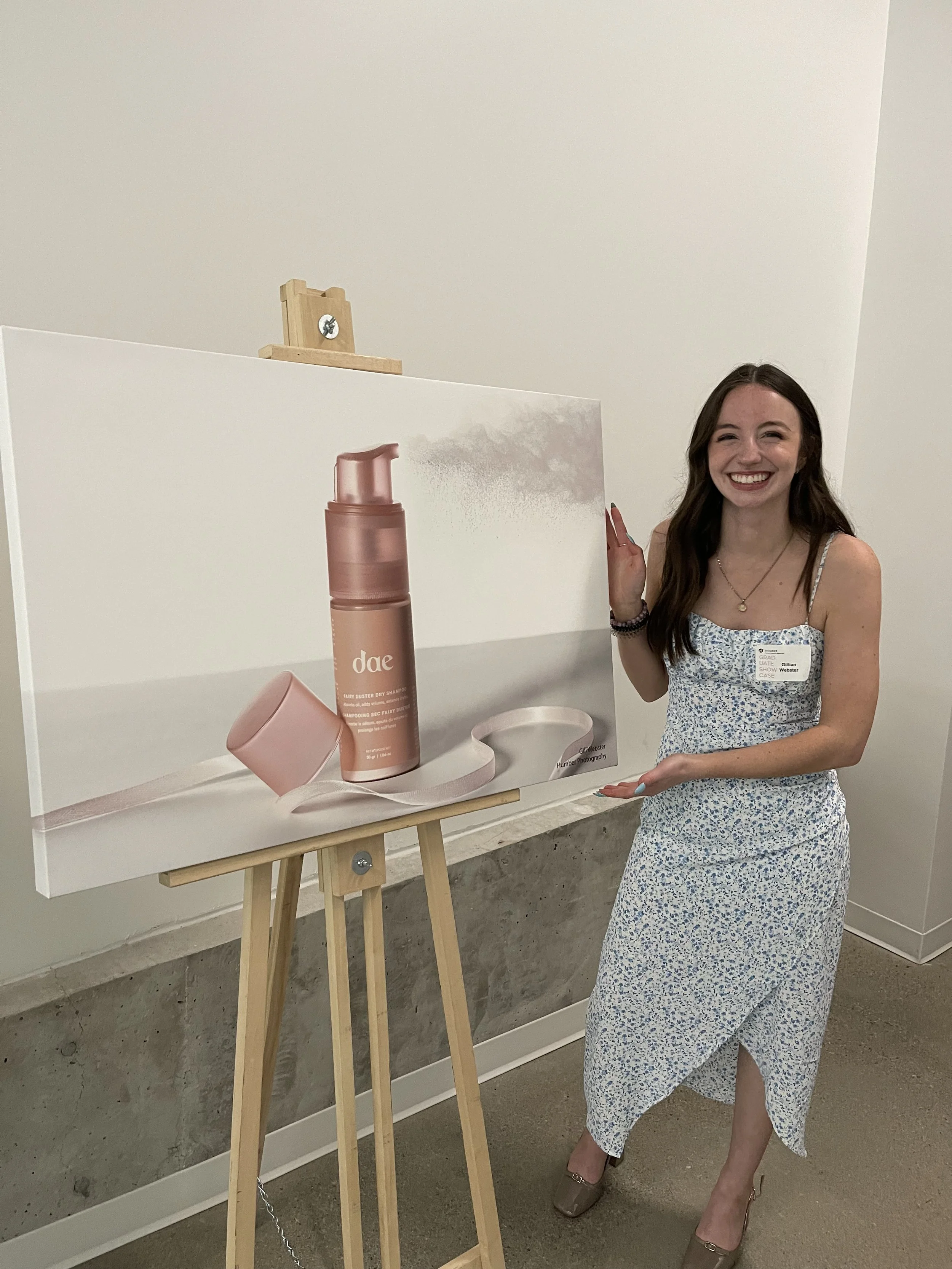 A woman wearing a floral dress stands next to a large poster on an easel. She is smiling and has long brown hair. The poster displays a pink bottle of spray, labeled "dae," with some text and a pink cup. The woman is wearing a name tag and beige high heels.