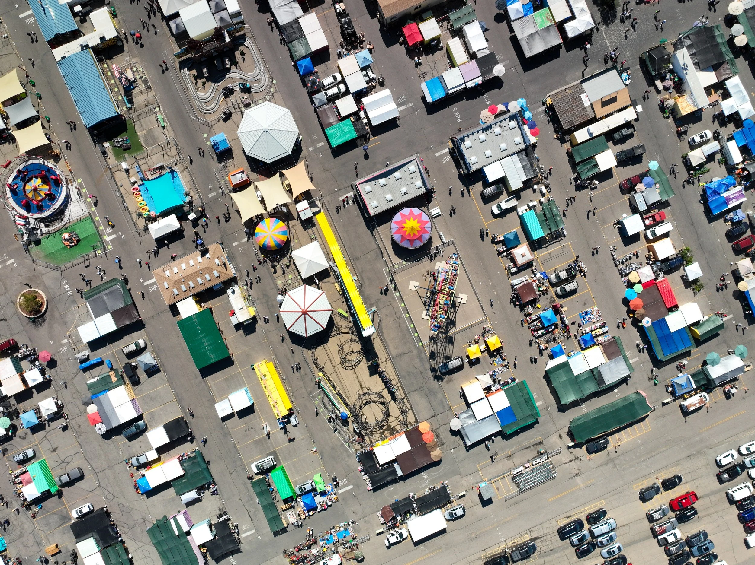 An aerial view of a busy outdoor fair with various booths, tents, and rides, filled with people and parked cars.
