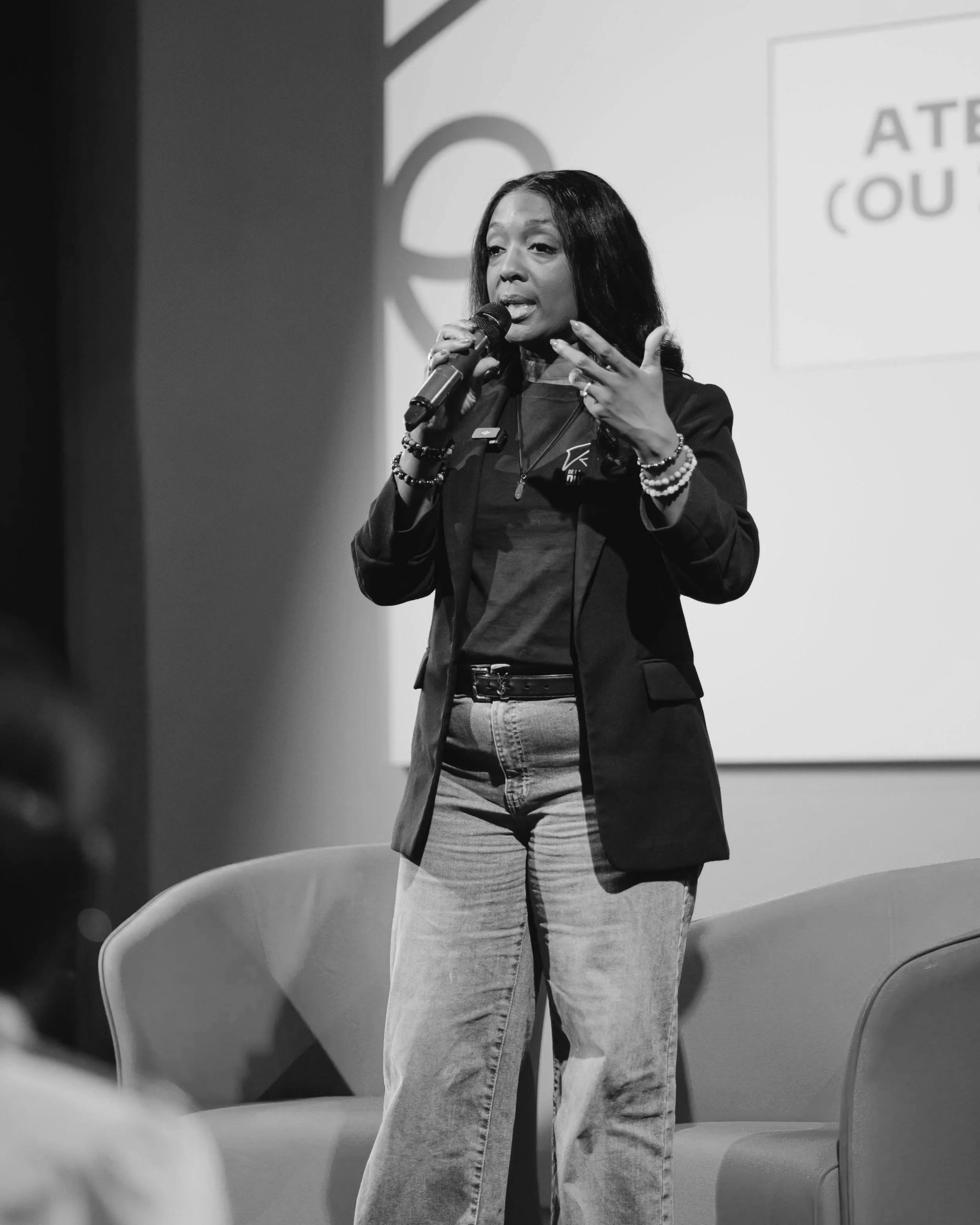 Une femme parle en public lors d'une conférence ou d'un événement, tenant un micro avec un écriteau derrière elle.