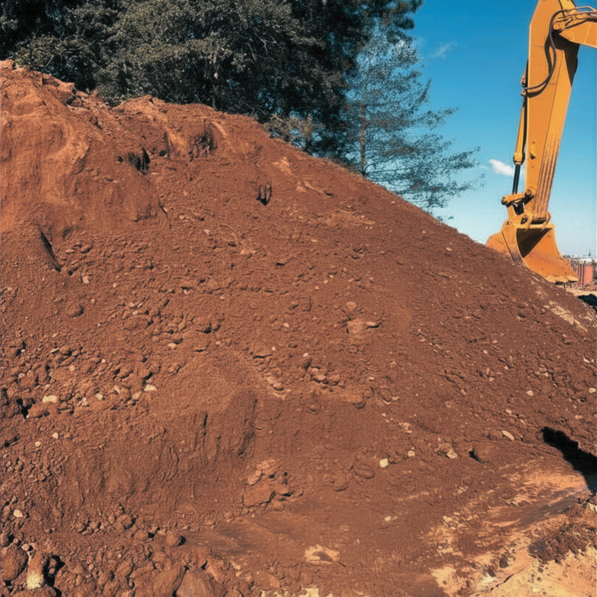 A construction site with a large pile of reddish soil and a yellow excavator arm visible on the right side, with a background of trees and a blue sky.