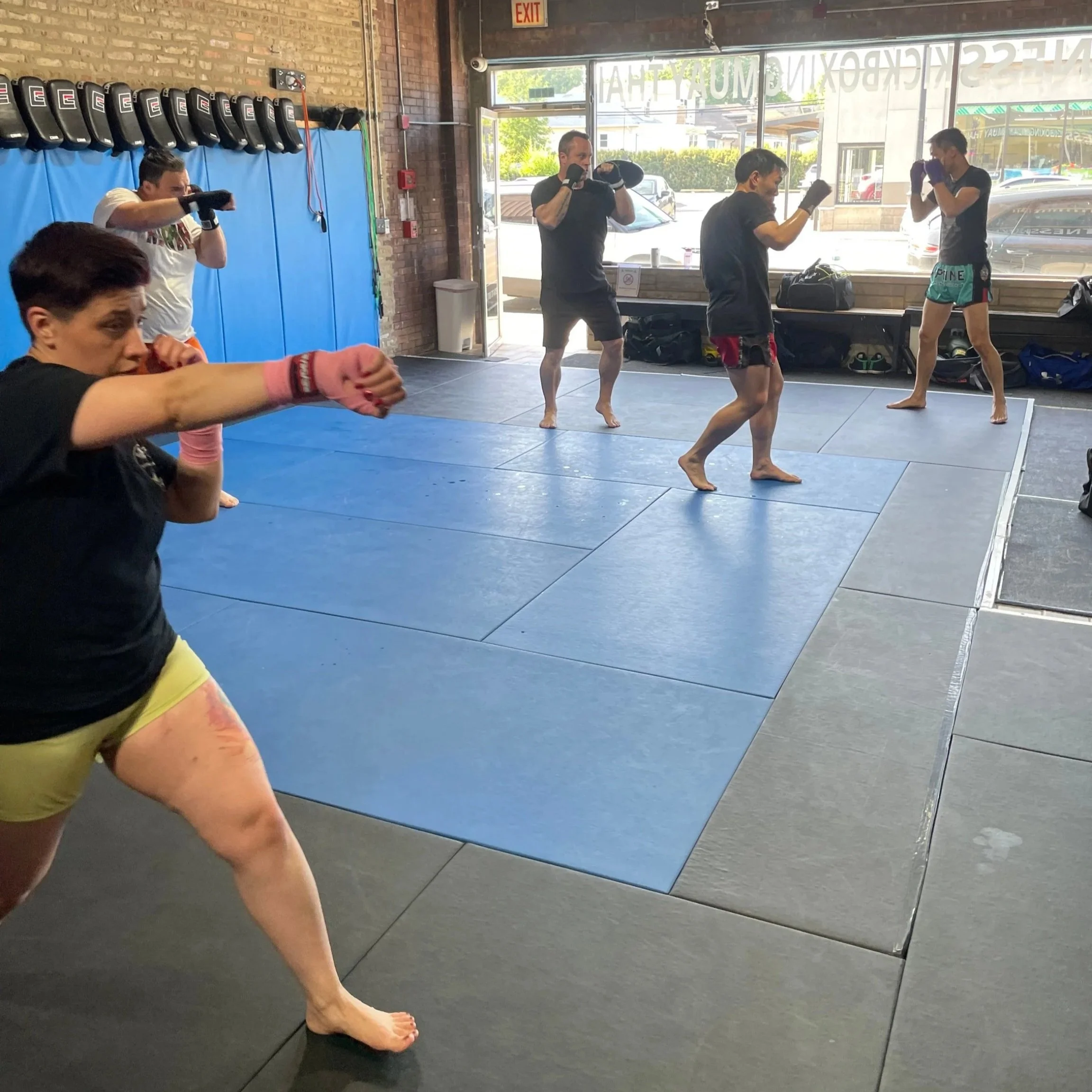 Group of people practicing Muay Thai or boxing in a gym with some wearing gloves and shorts, in a stance ready to punch or defend.