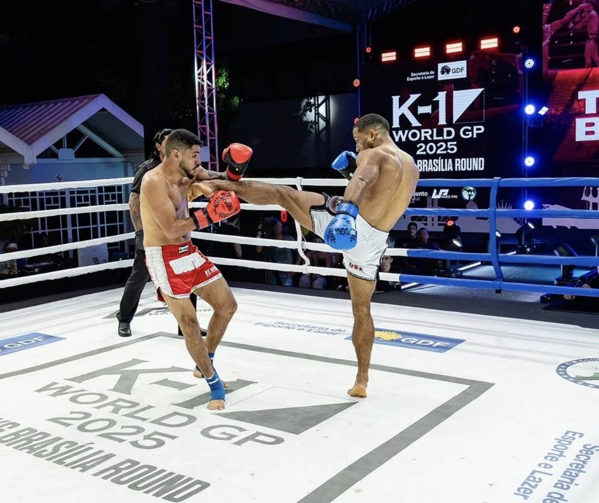 A muay Thai or kickboxing match with two fighters in the ring, one delivering a high kick to the other. The fighter with red shorts and gloves is receiving the kick from the fighter in white shorts and blue gloves. An official stands behind them, observing. The ring has branding for the K-1 World GP 2025 event, with the location noted as Brasilia.