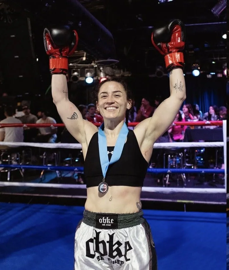 A female kickboxer in a black sports bra and boxing shorts, wearing red boxing gloves and a medal around her neck, smiling with arms raised in a boxing ring.