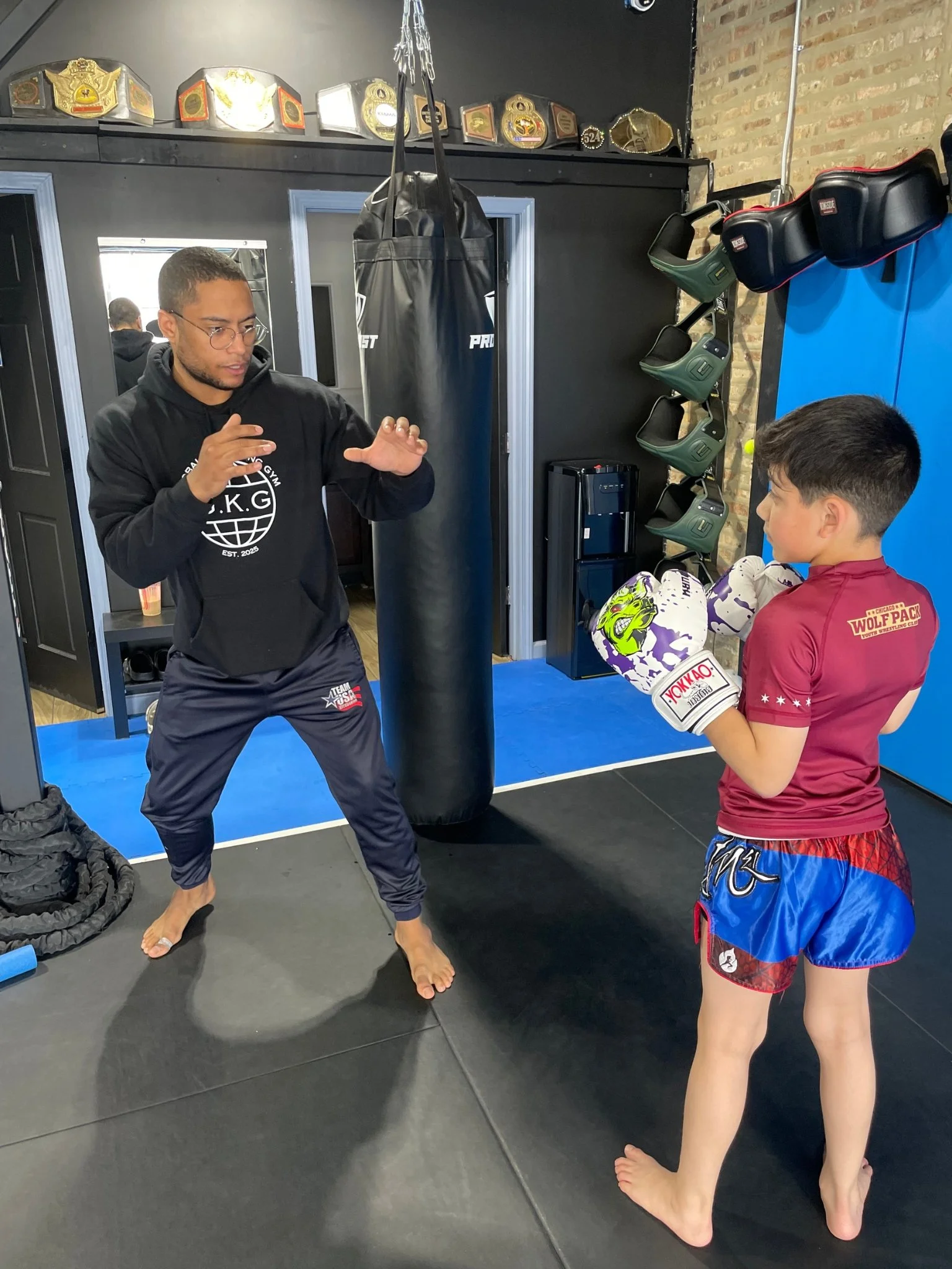 A man in black gym attire demonstrating Kickboxing and Muay Thai technique to a young boy in boxing gloves. They are standing inside a gym with boxing gear, championship belts, and training equipment on the walls.
