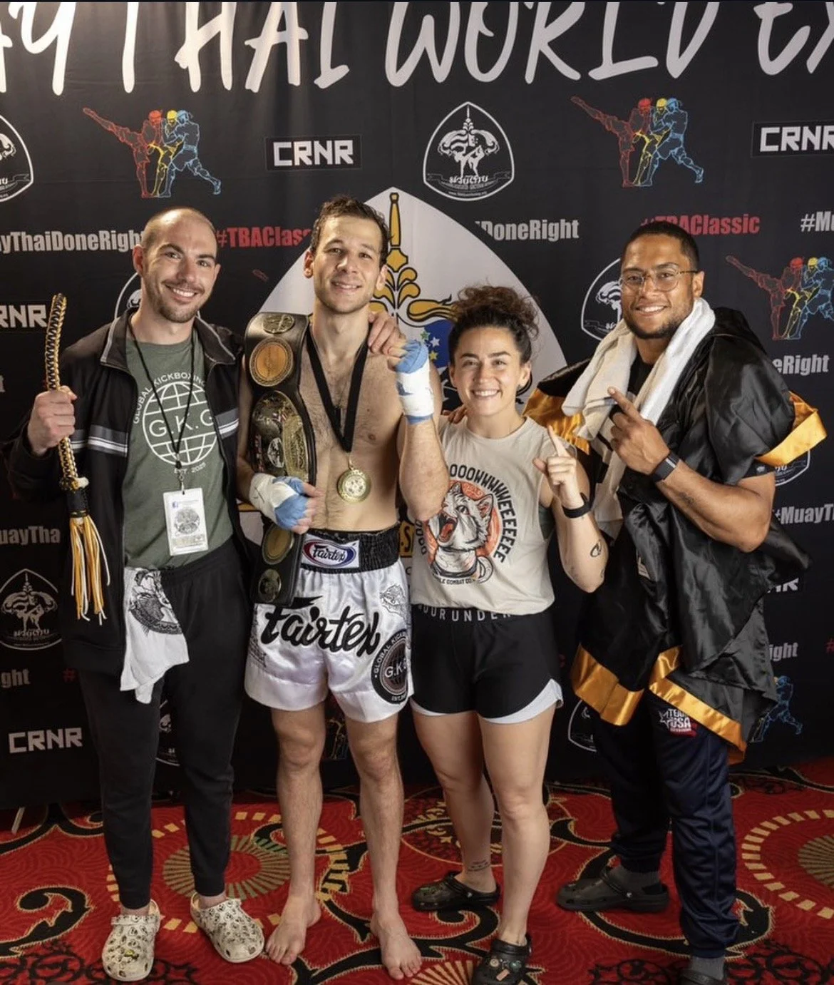 A group of four people celebrating a martial arts victory at a competition. The group includes a shirtless man with medals and a championship belt, a man on the left wearing a jacket and lanyard, a woman wearing sports shorts and a T-shirt, and a man on the right wearing a cape and pointing. They are standing in front of a black backdrop with martial arts logos and text.