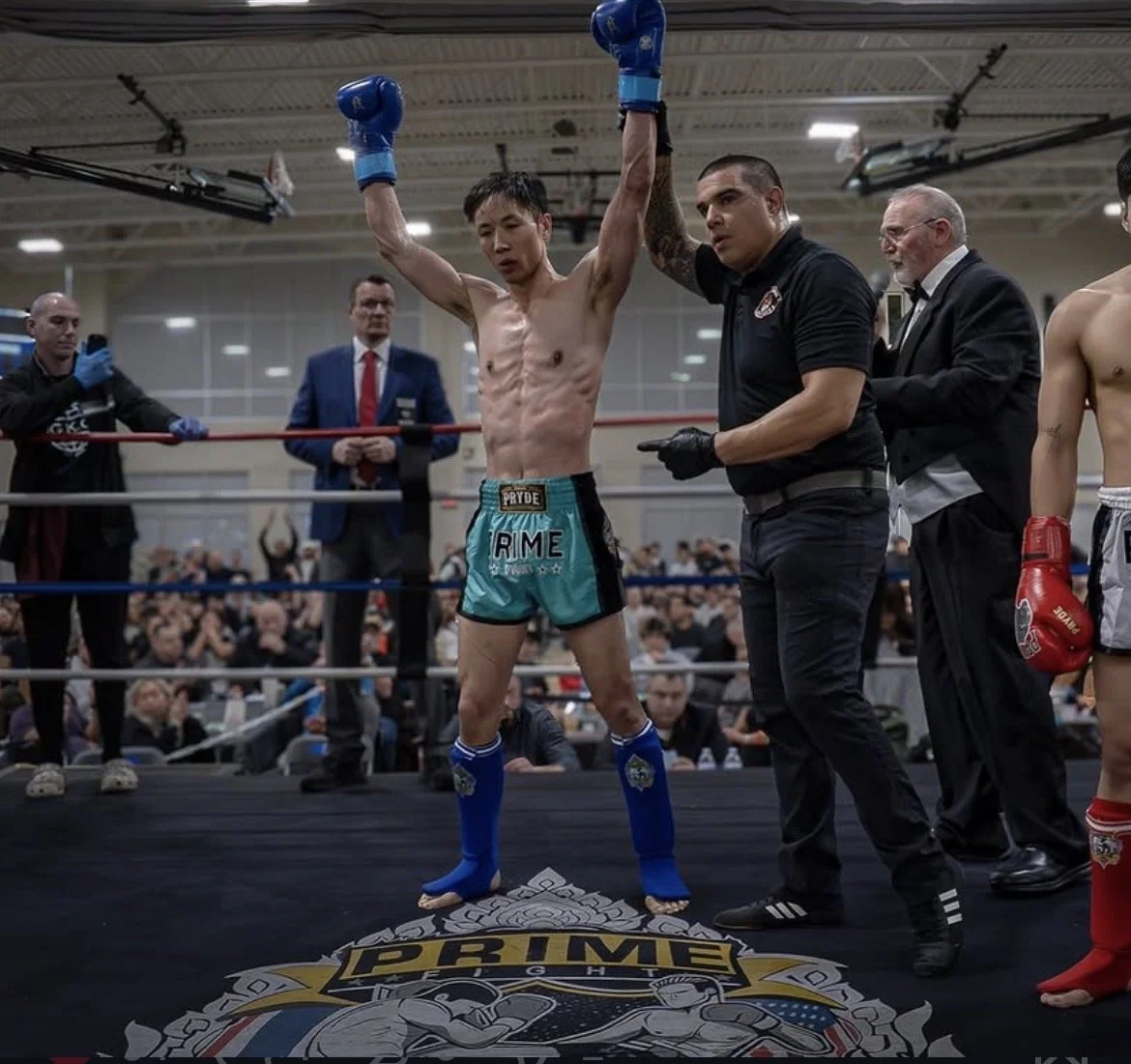 A victorious male Thai boxer with arms raised in the ring, surrounded by officials and fellow fighters, in front of a crowd and a prize logo on the floor.