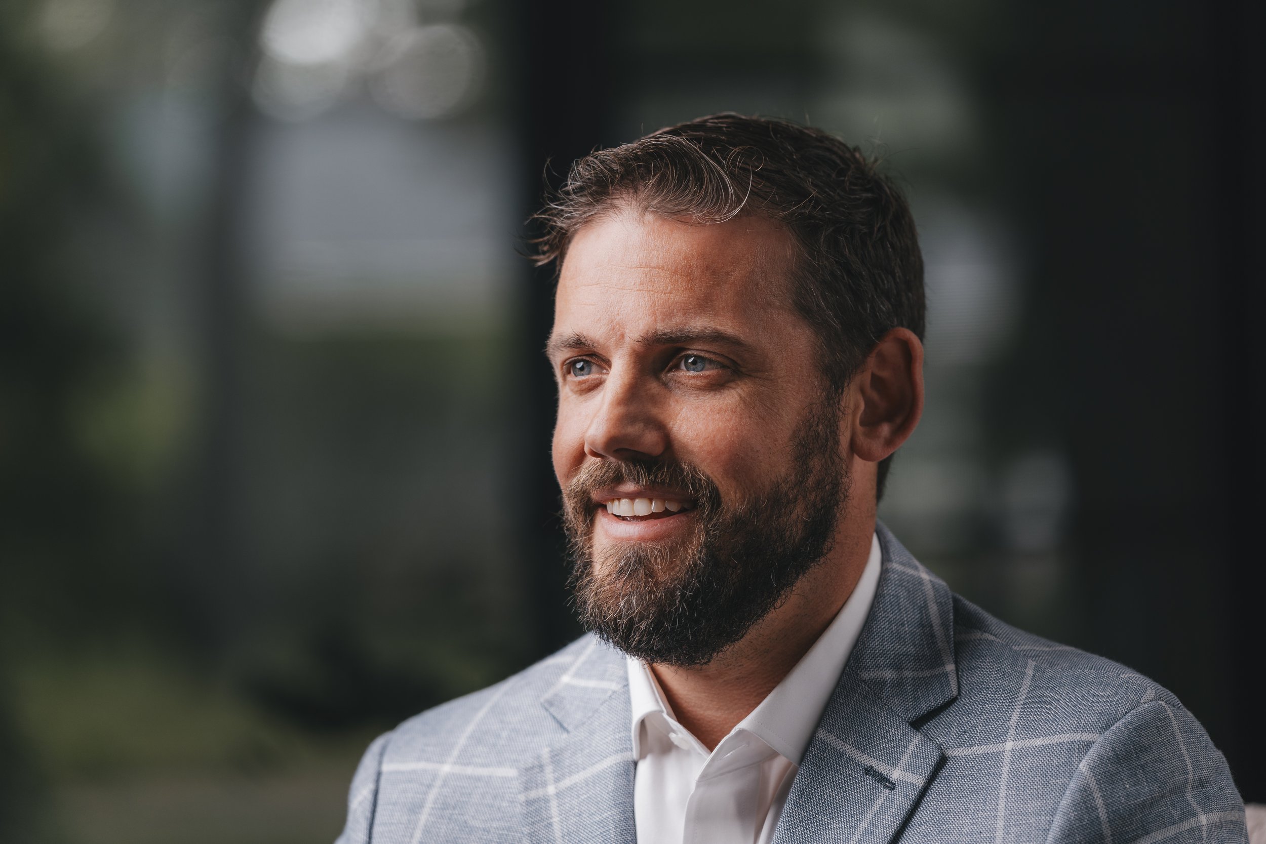 Close-up of a smiling man with dark hair and a beard, wearing a light-colored plaid blazer and white shirt, outdoors with blurred greenery background.