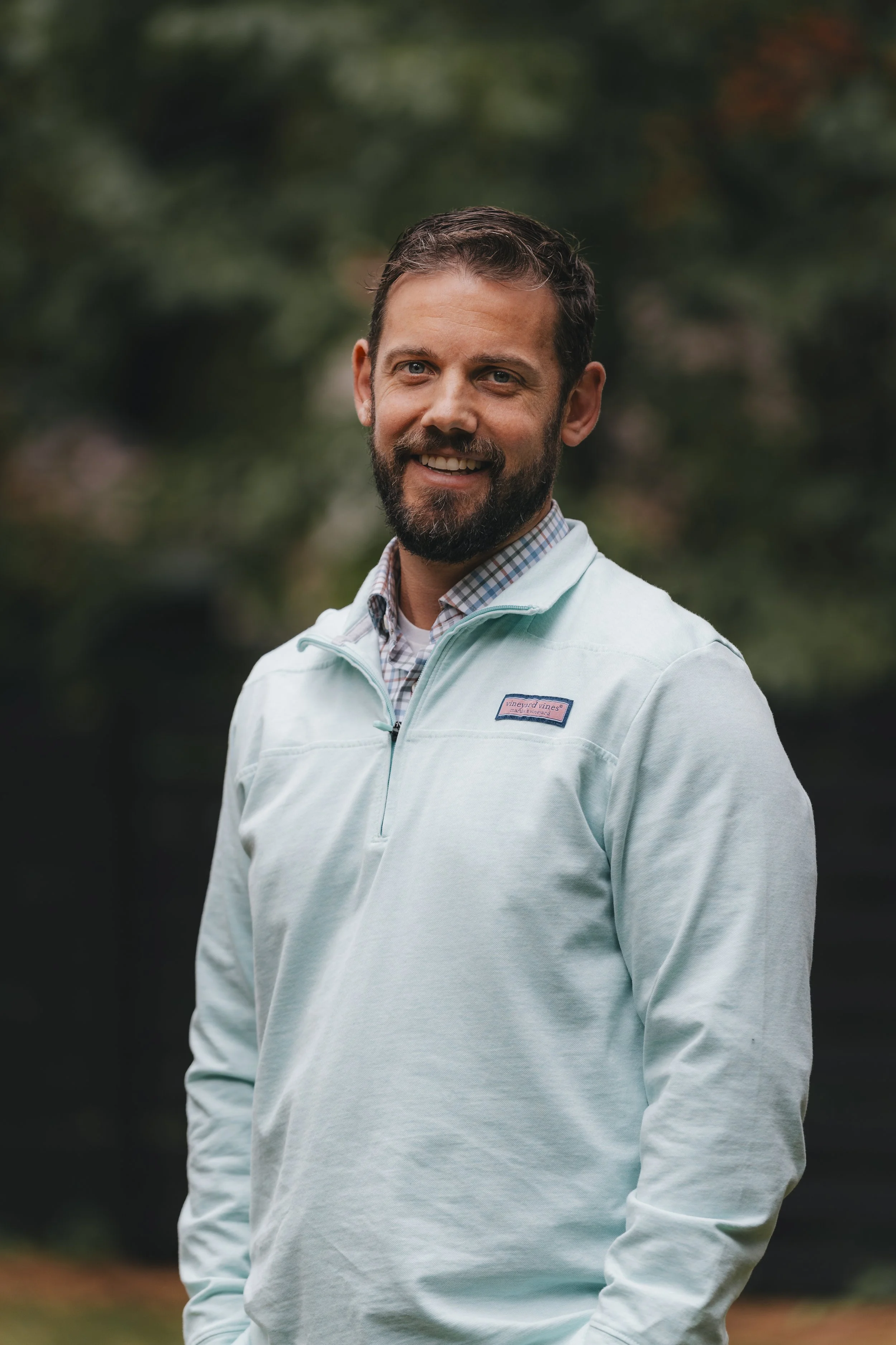 A smiling man with a beard and short dark hair, wearing a light blue quarter-zip pullover over a collared shirt, standing outdoors with a blurred green and orange background.