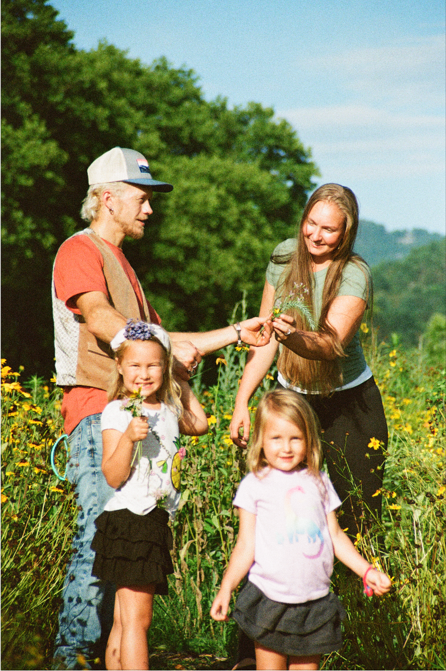 A family enjoying a walk through a yellow wildflower field on a sunny day, with two young girls in the foreground and two adults behind them.