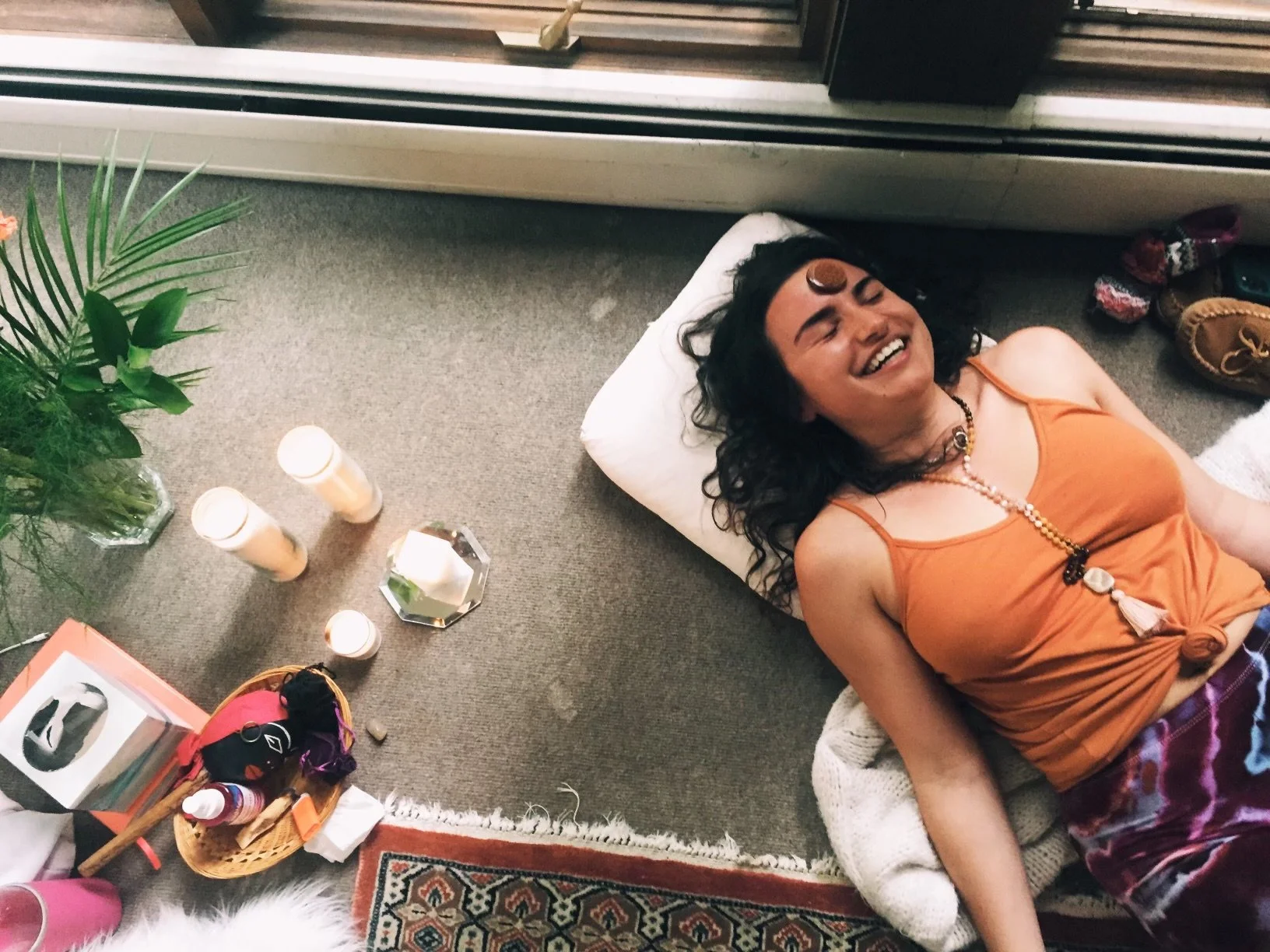 A woman lying on the floor, smiling and laughing with a gemstone on her forehead, surrounded by candles, plants, and various objects.