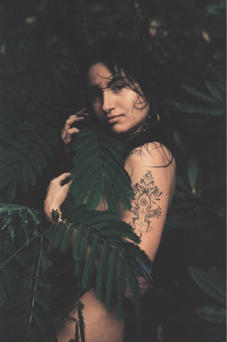 A woman with wet hair posing among green forest foliage, showing a tattoo on her arm.