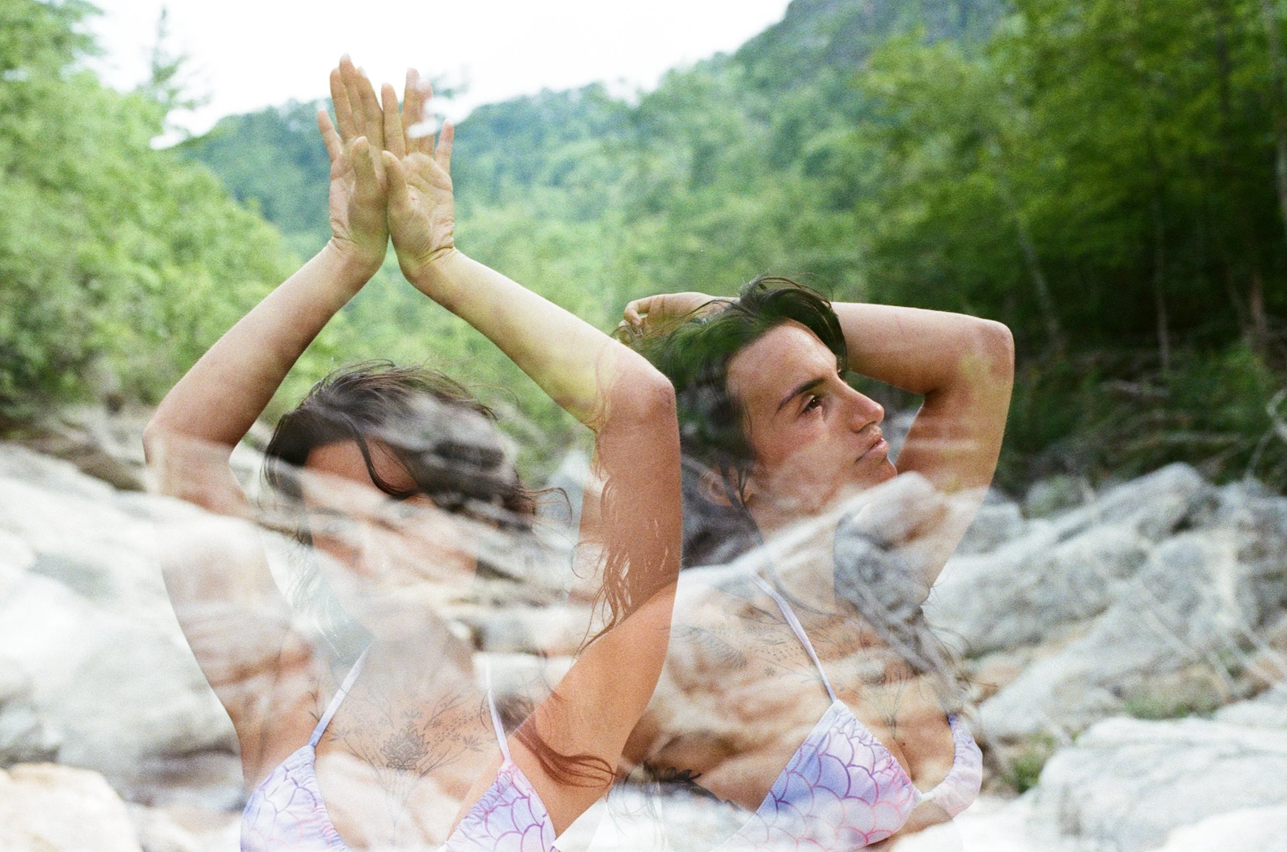 Two women wearing swimsuits are practicing yoga outdoors near rocks and a river, surrounded by green trees.