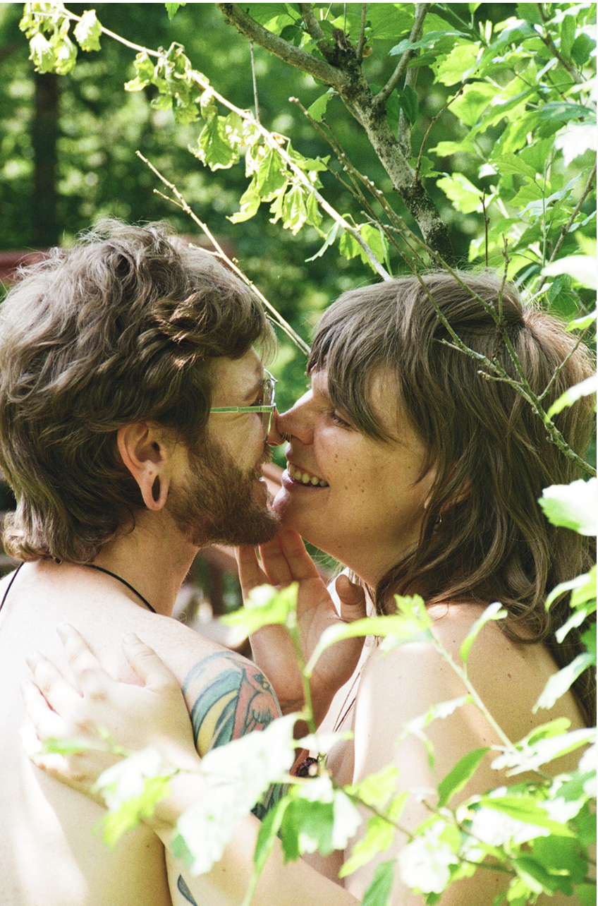 Two people with short hair and tattoos smiling and touching noses amidst green foliage.