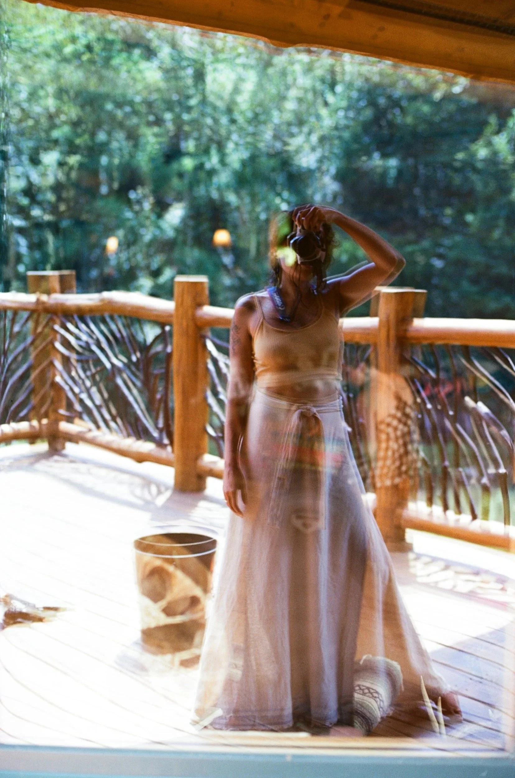 A woman standing on a wooden deck outdoors, seen through a glass window, with trees in the background. She is wearing a beige tank top and a long, sheer skirt, with her hand on her head.