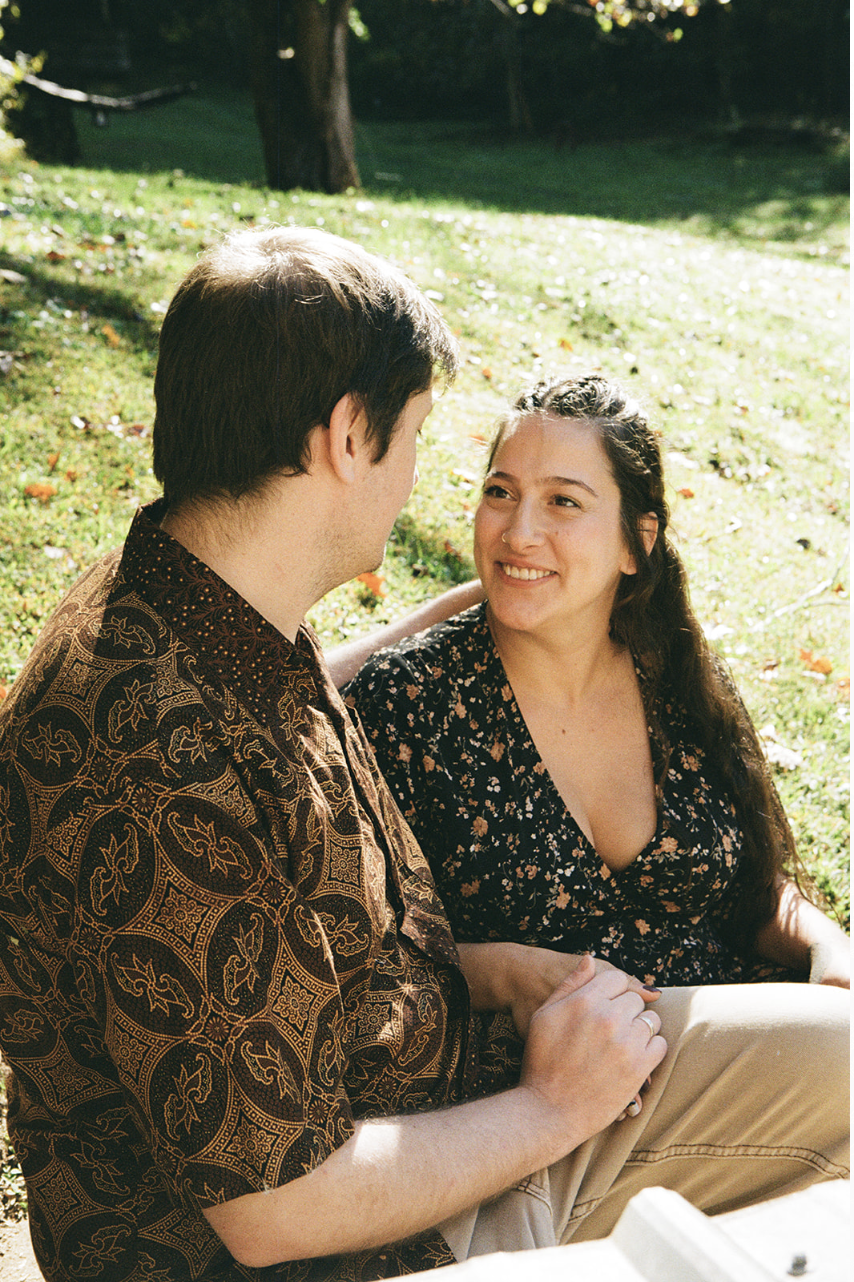 A man and woman are sitting outdoors on grass, engaging in a conversation, with trees in the background, during daytime.